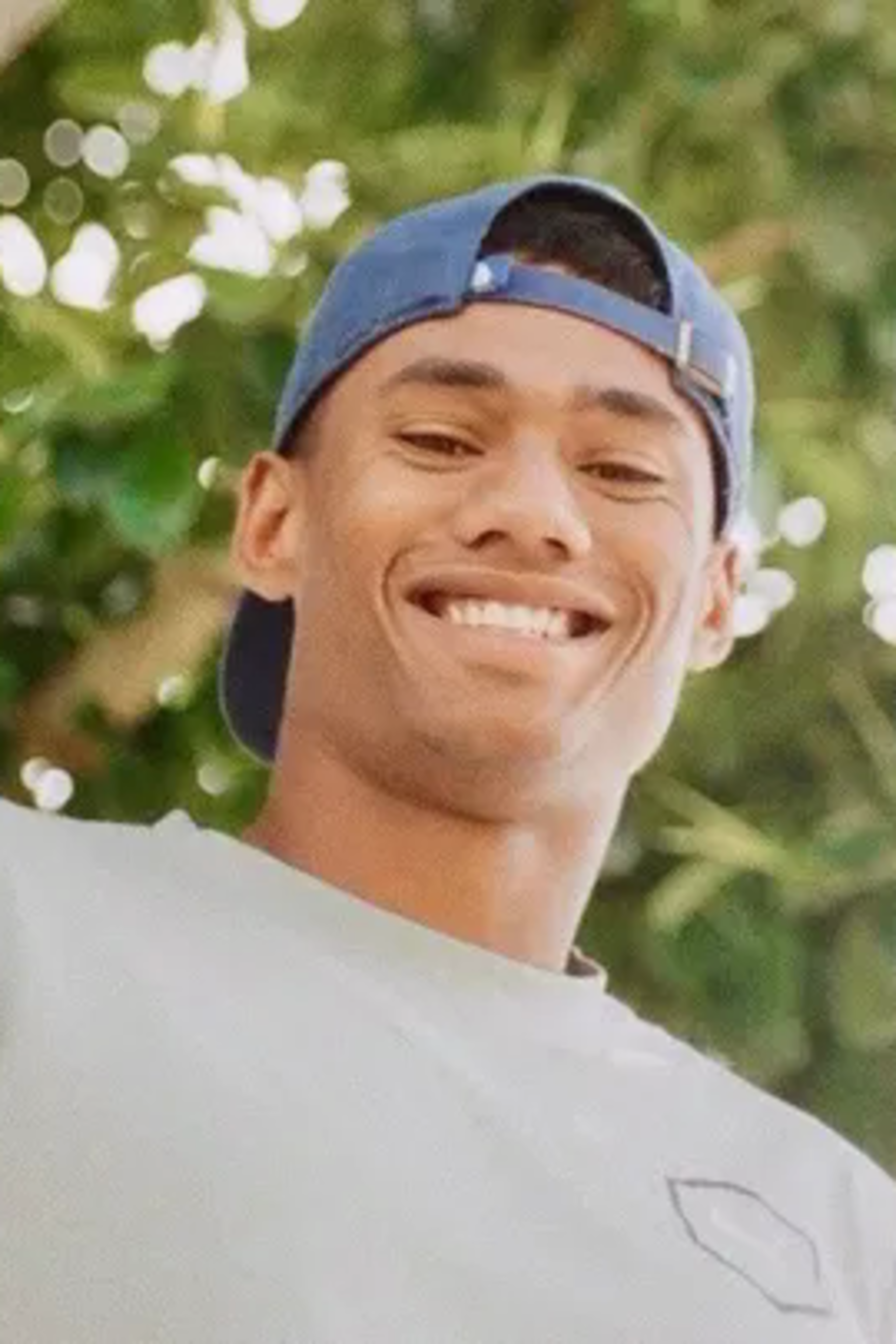 A young man wearing a blue backward cap and a light gray shirt smiles warmly with greenery and bright sunlight in the background.