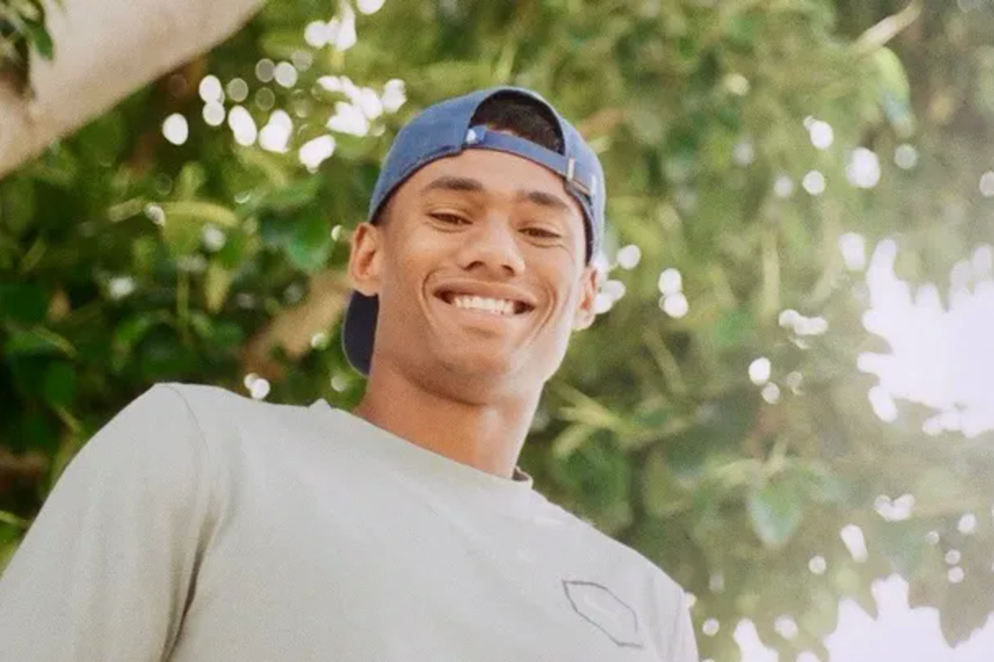 A young man wearing a blue backward cap and a light gray shirt smiles warmly with greenery and bright sunlight in the background.