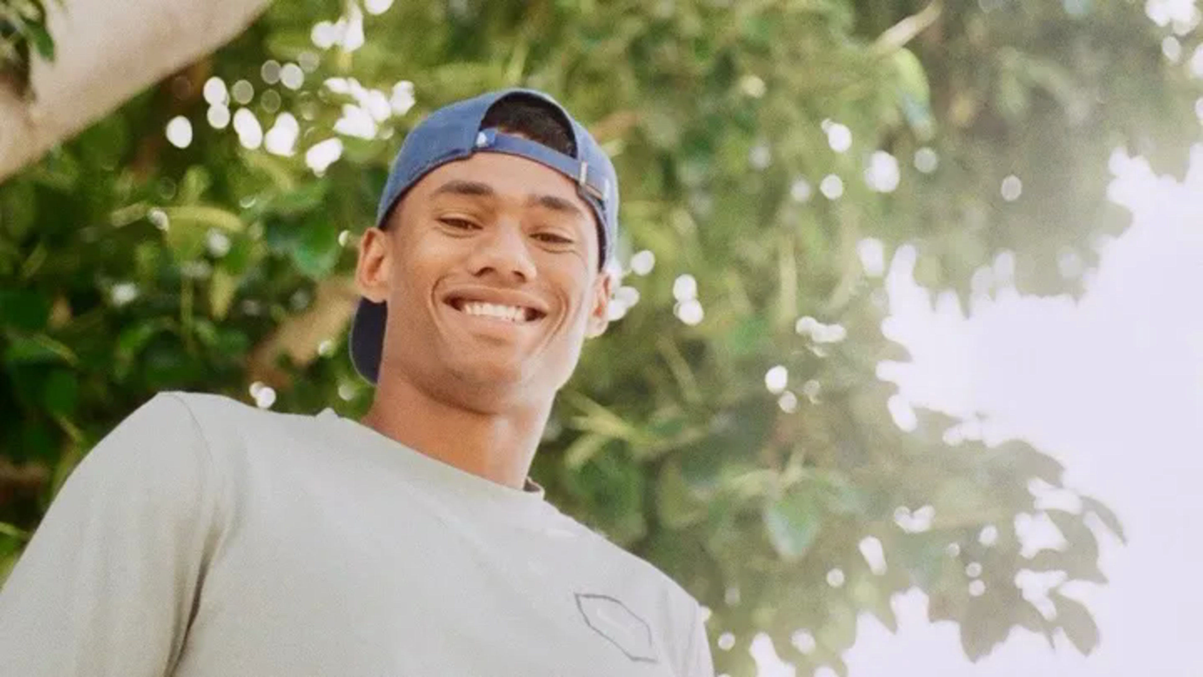 A young man wearing a blue backward cap and a light gray shirt smiles warmly with greenery and bright sunlight in the background.
