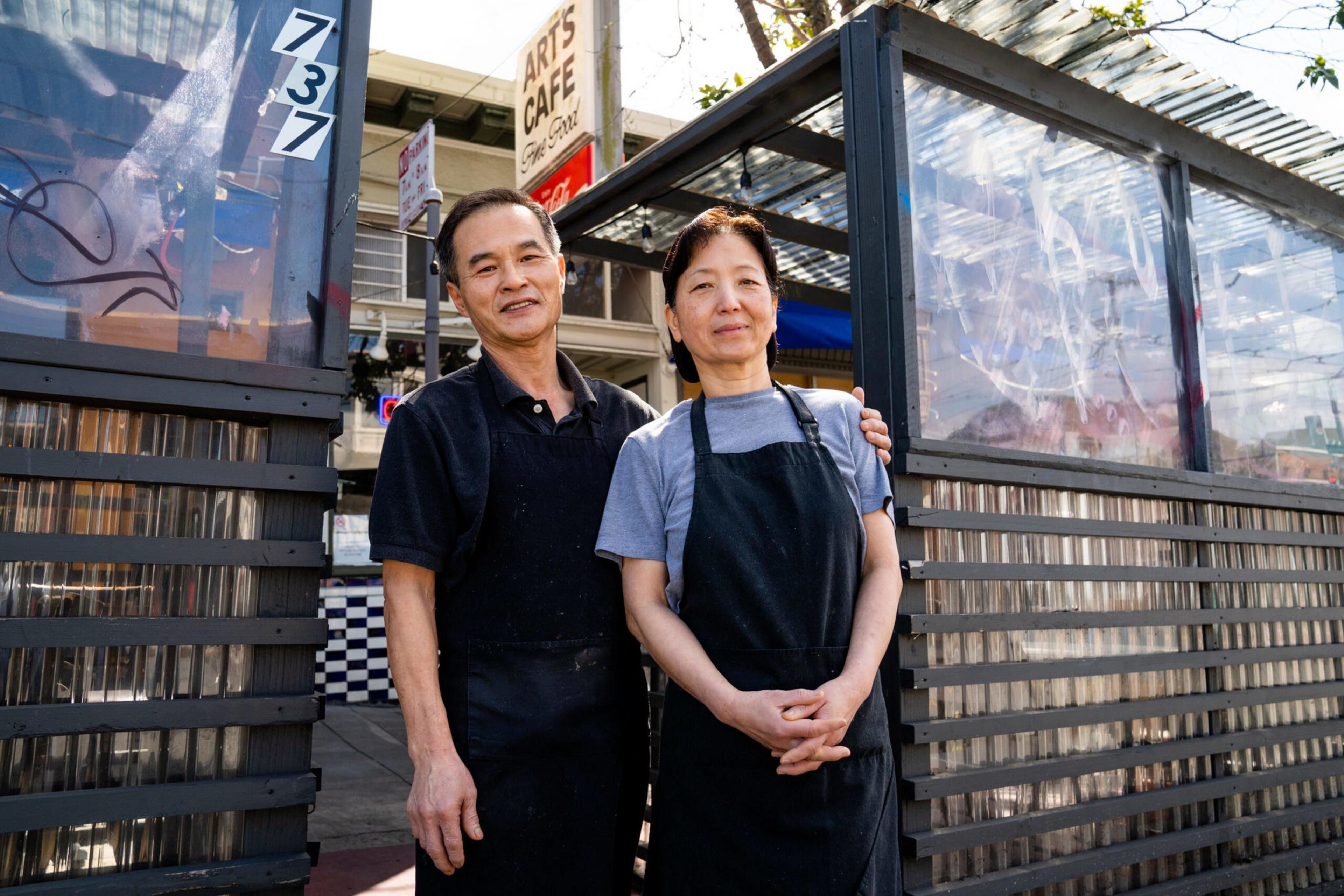 A man and woman in black aprons stand outside a café, smiling. The café sign reads &quot;Art's Cafe.&quot; They appear to be in a sunny urban setting.