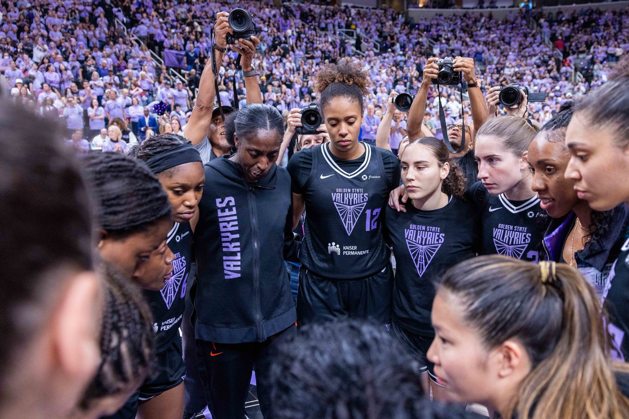 A women's basketball team wearing Golden State Valkyries jerseys is huddled closely together, with intense focus and a crowd behind them.