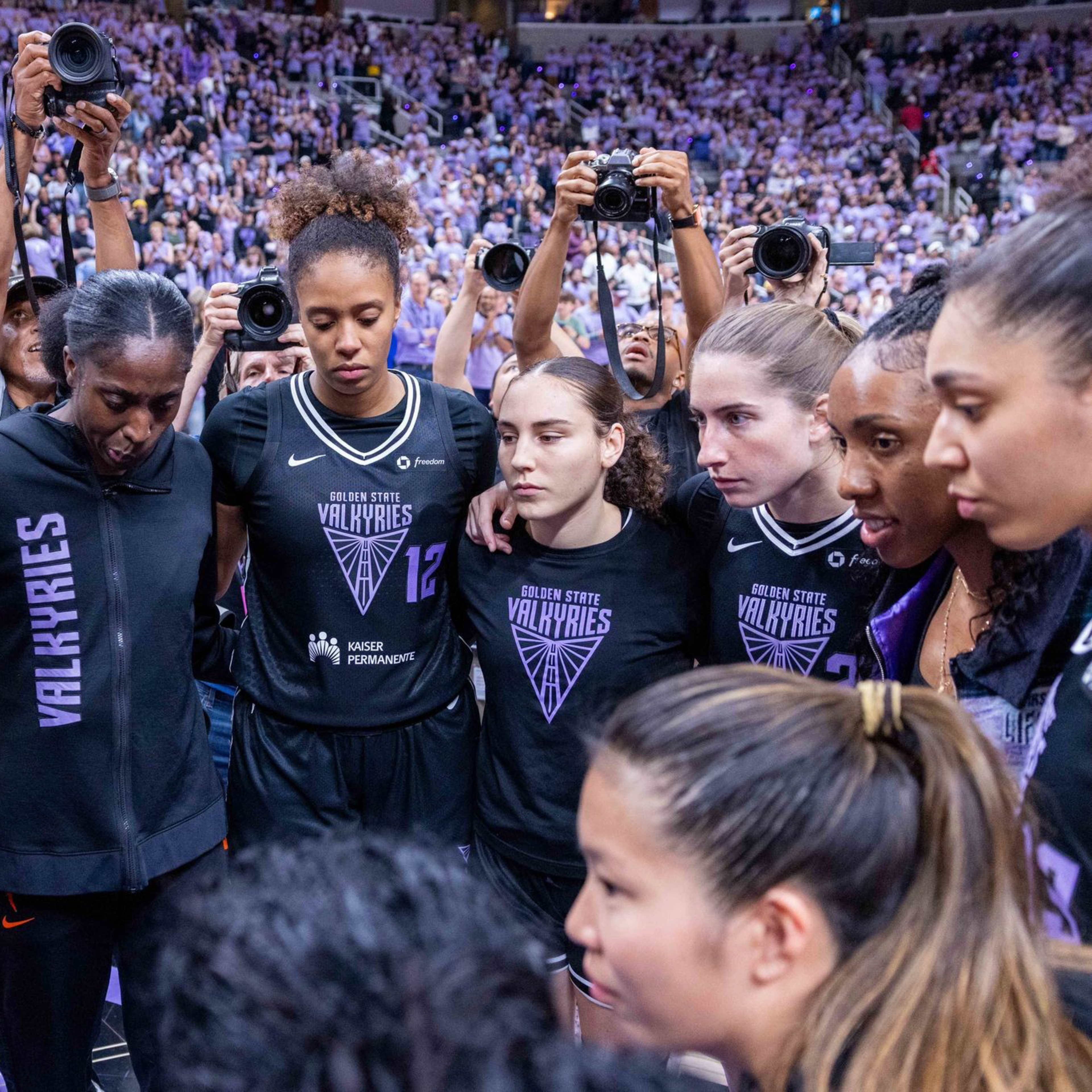 A women's basketball team wearing Golden State Valkyries jerseys is huddled closely together, with intense focus and a crowd behind them.