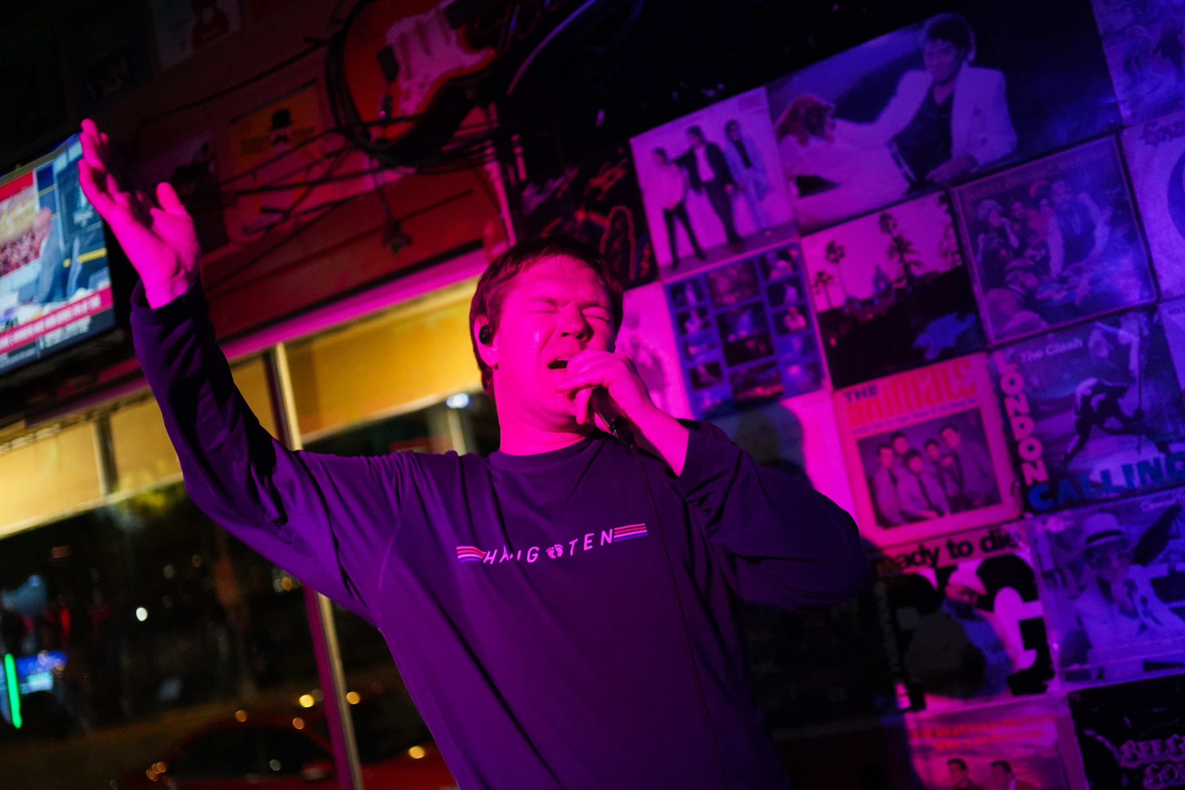 A person passionately sings into a microphone under purple lighting, with one arm raised, in a room decorated with vintage music posters and albums.