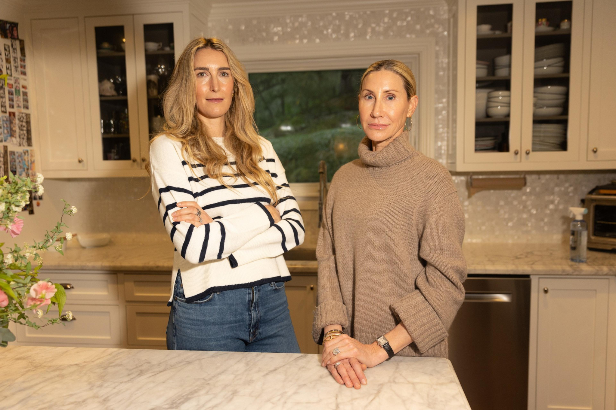 Two women stand in a kitchen with cream cabinets and marble countertops. One wears a striped sweater; the other, a tan turtleneck. Flowers are visible nearby.