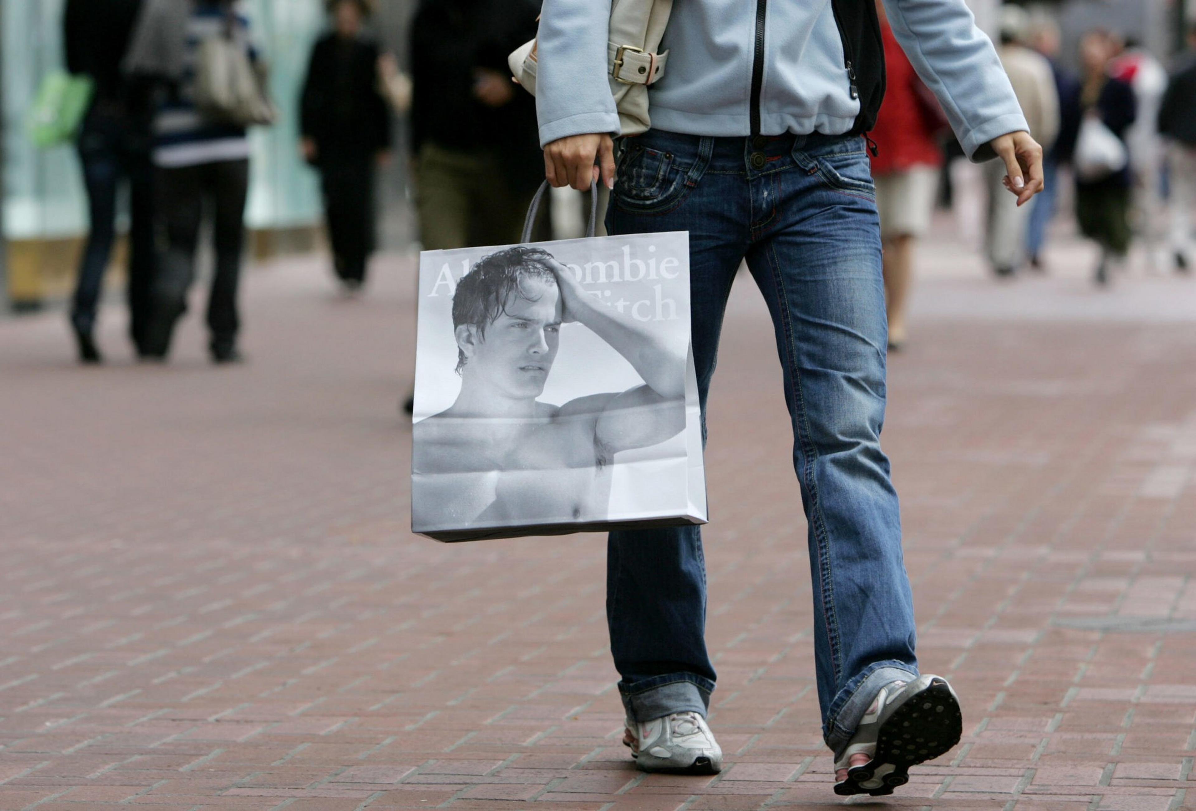 A person in jeans and sneakers walks on a brick sidewalk, holding a black-and-white Abercrombie & Fitch shopping bag with a shirtless male model pictured.