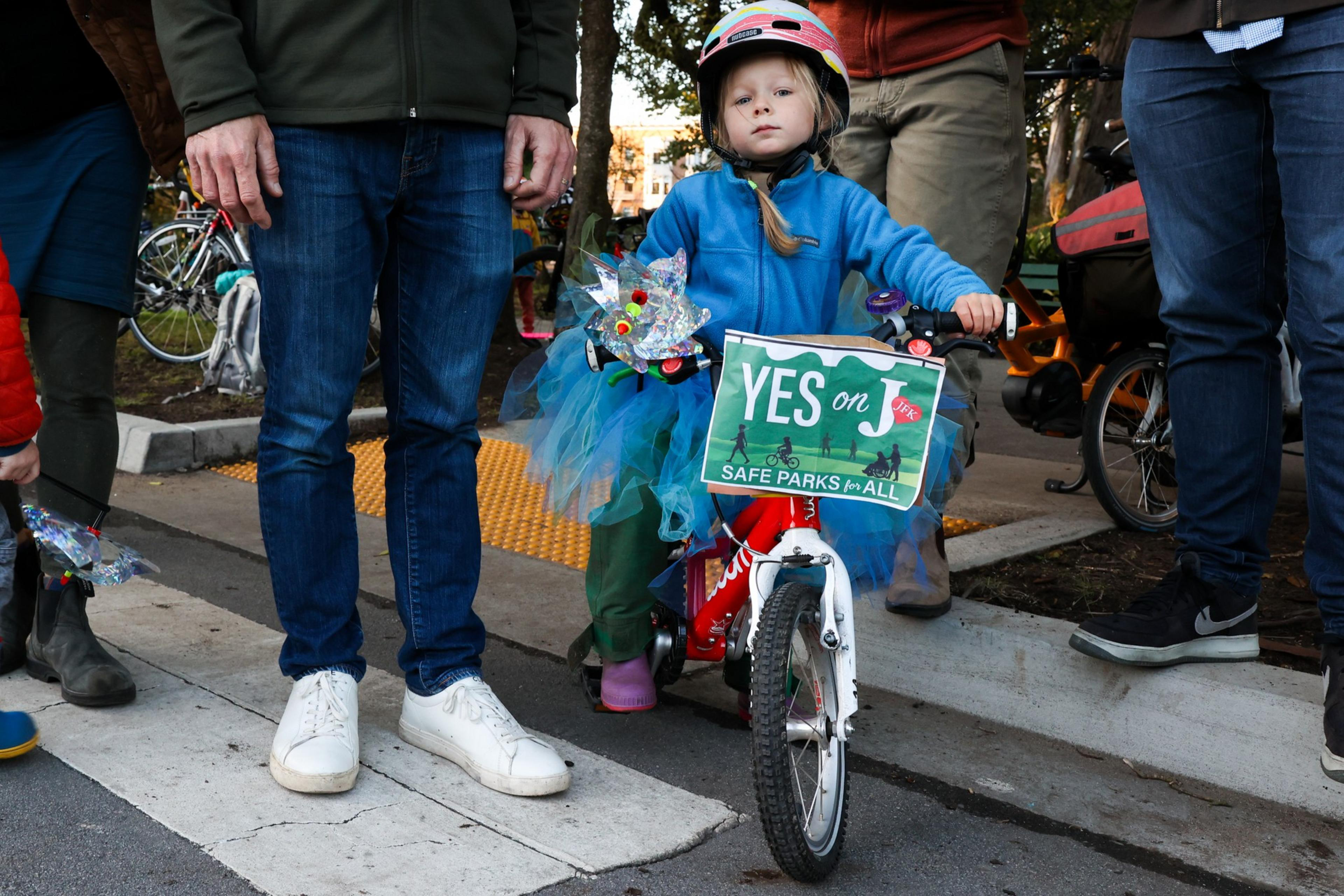 A young child wearing a blue jacket and pink helmet sits on a red bike with a "YES on J" sign; adults stand nearby on a street with trees in the background.