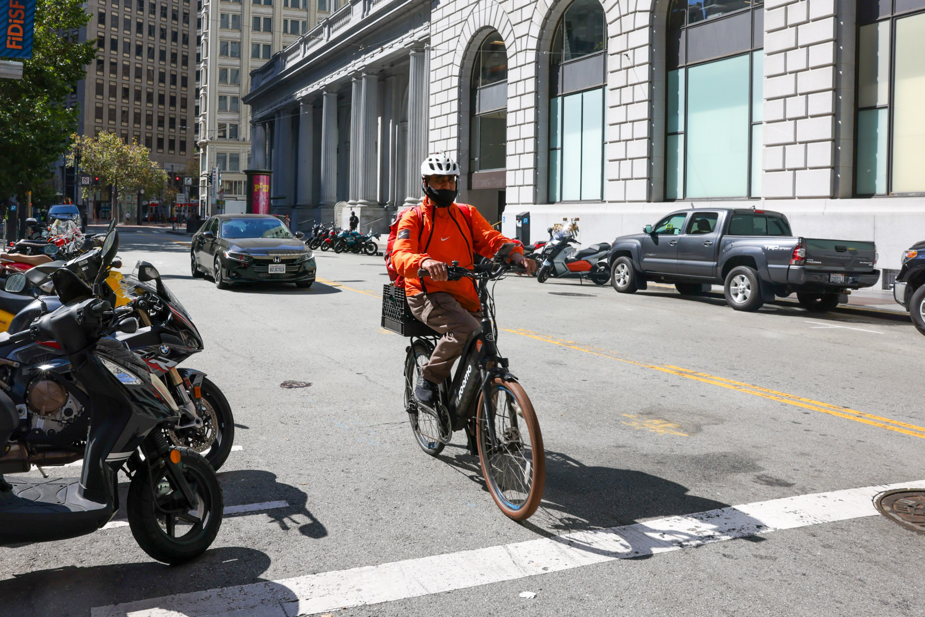 A person wearing an orange jacket and white helmet rides a black electric bike on a city street lined with parked cars and scooters.