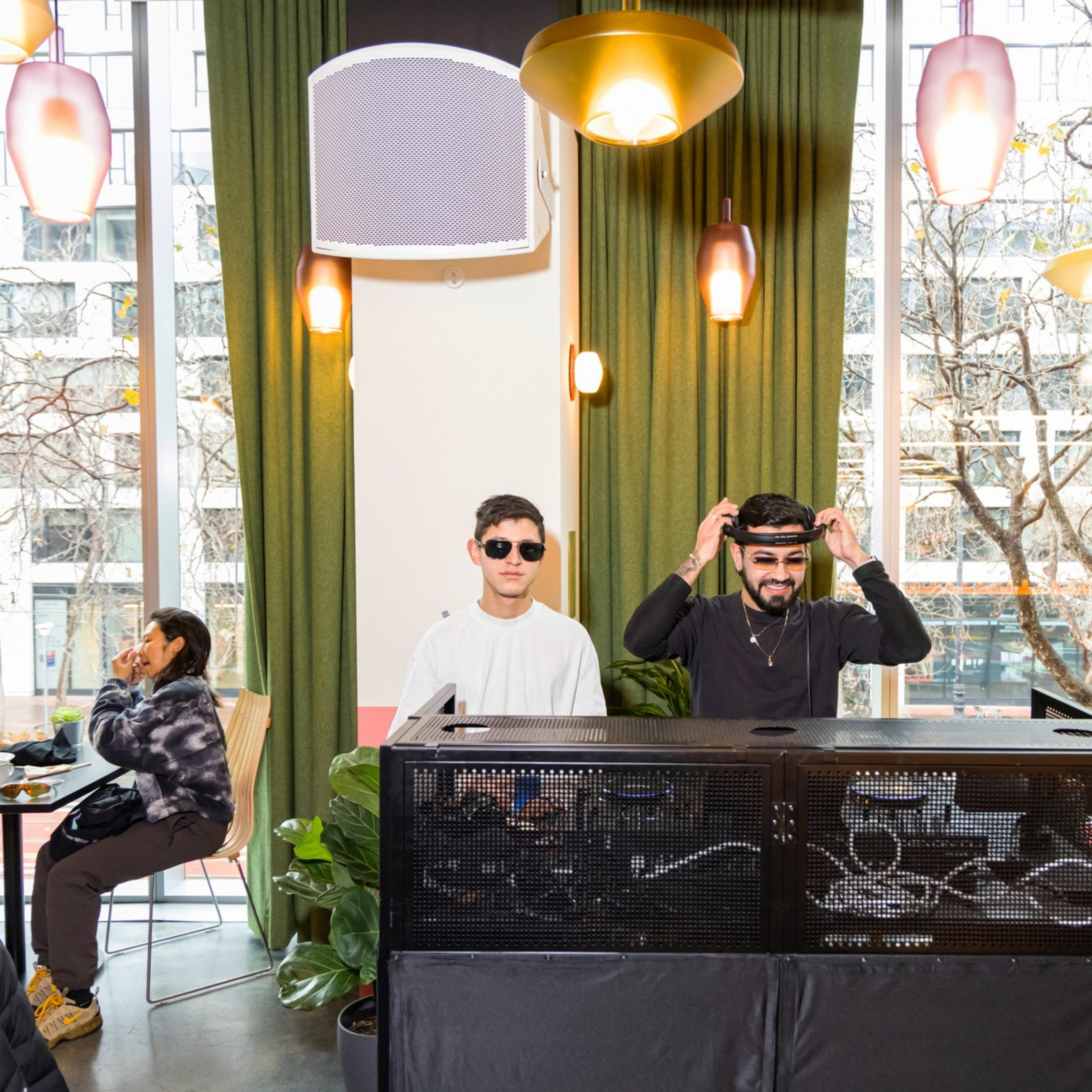 Two men stand behind DJ equipment in a cafe with large windows. One adjusts headphones, the other wears sunglasses. Diners are seated at tables nearby.