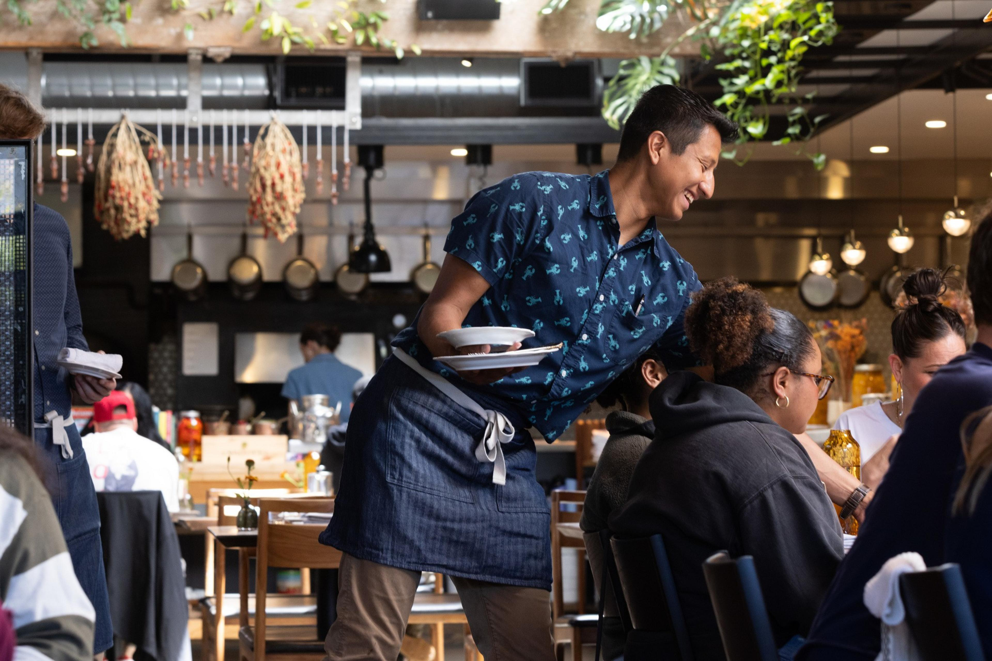 A waiter in a patterned shirt and apron smiles as he serves food to a group of people seated in a lively restaurant with hanging plants and a busy kitchen.