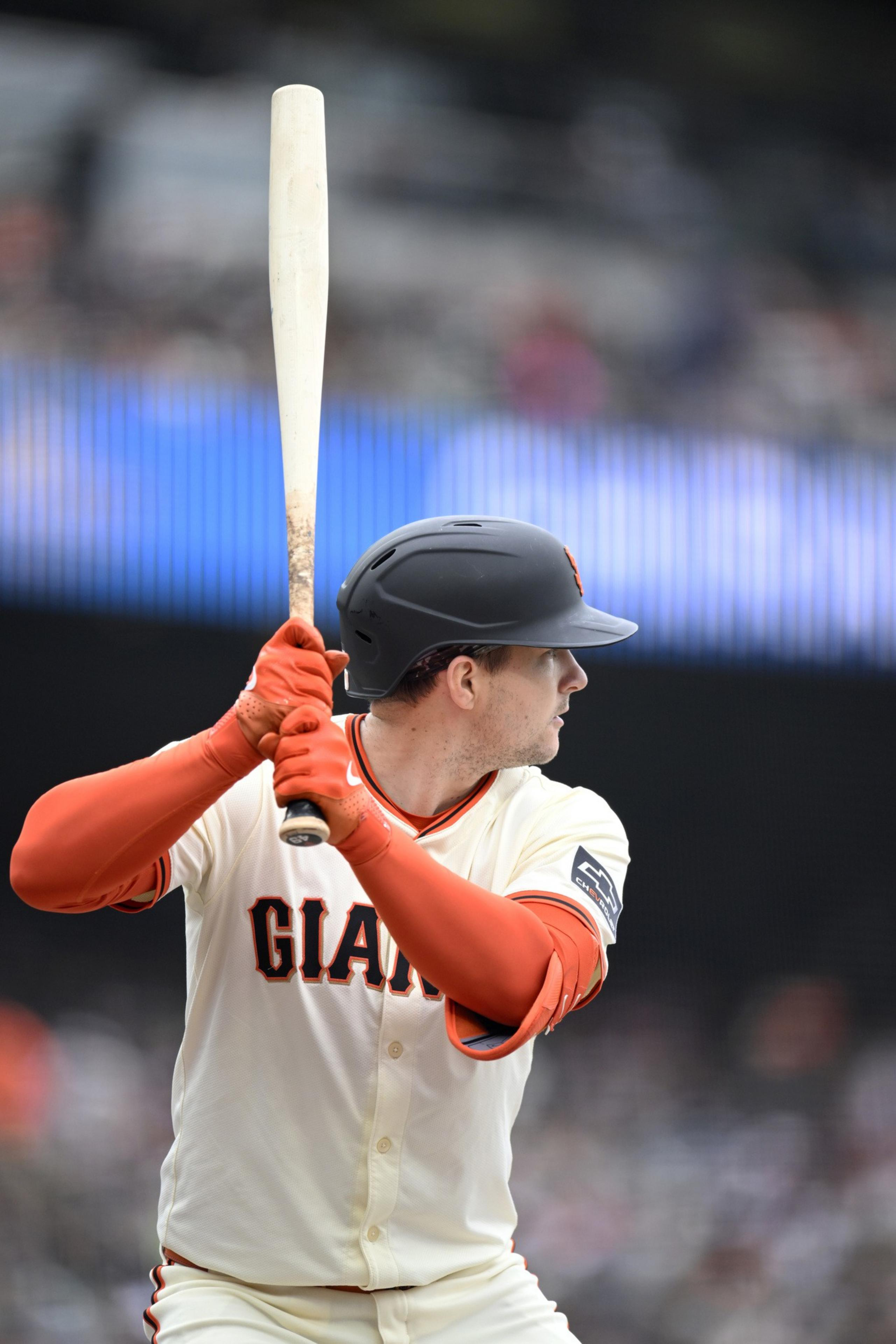 A baseball player in a white &quot;Giants&quot; jersey and black helmet is poised to bat, holding a light-colored bat, with a blurred stadium background.