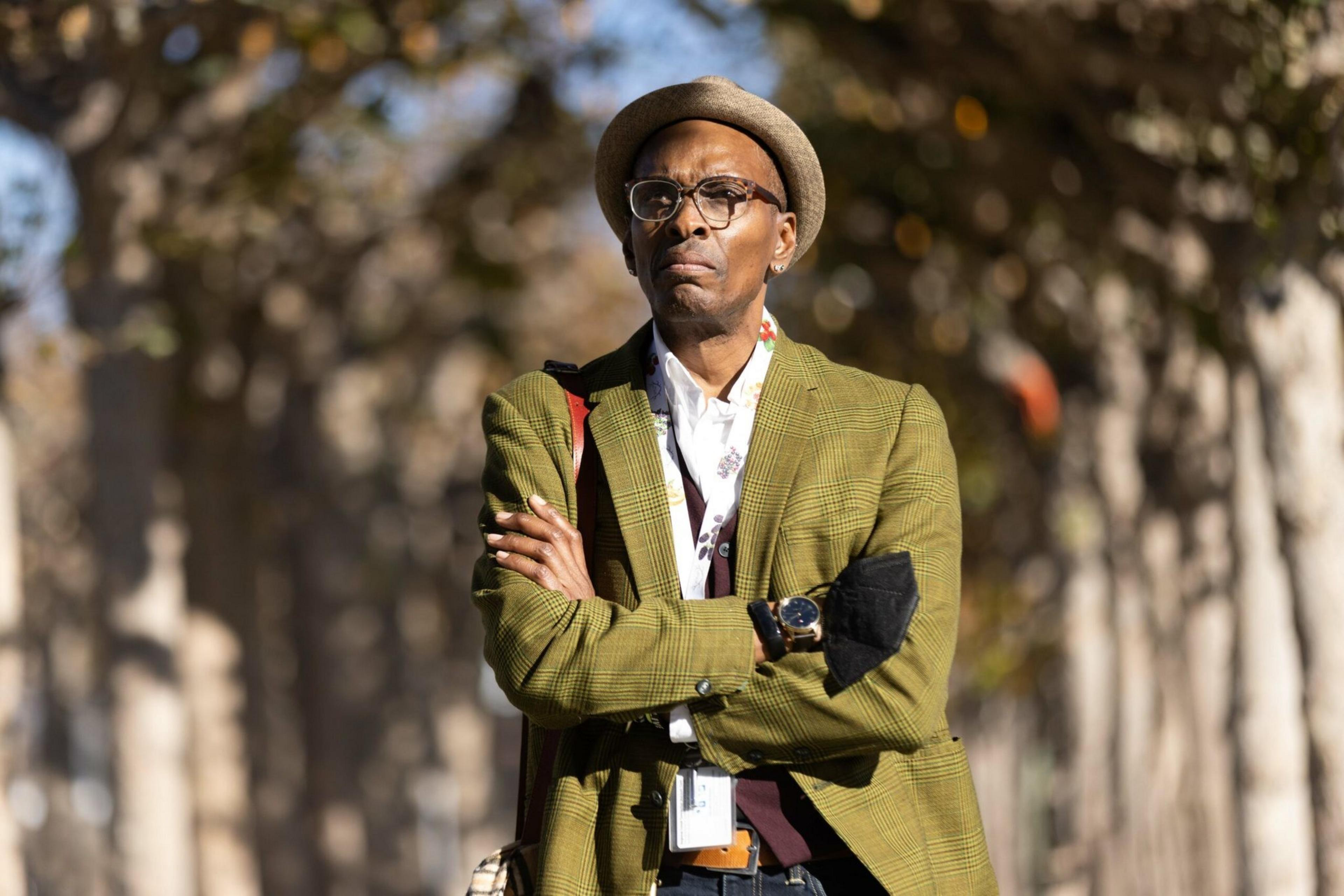 A man wearing glasses, a brown hat, and a green plaid blazer stands outdoors with his arms crossed, against a blurred background of tall trees.