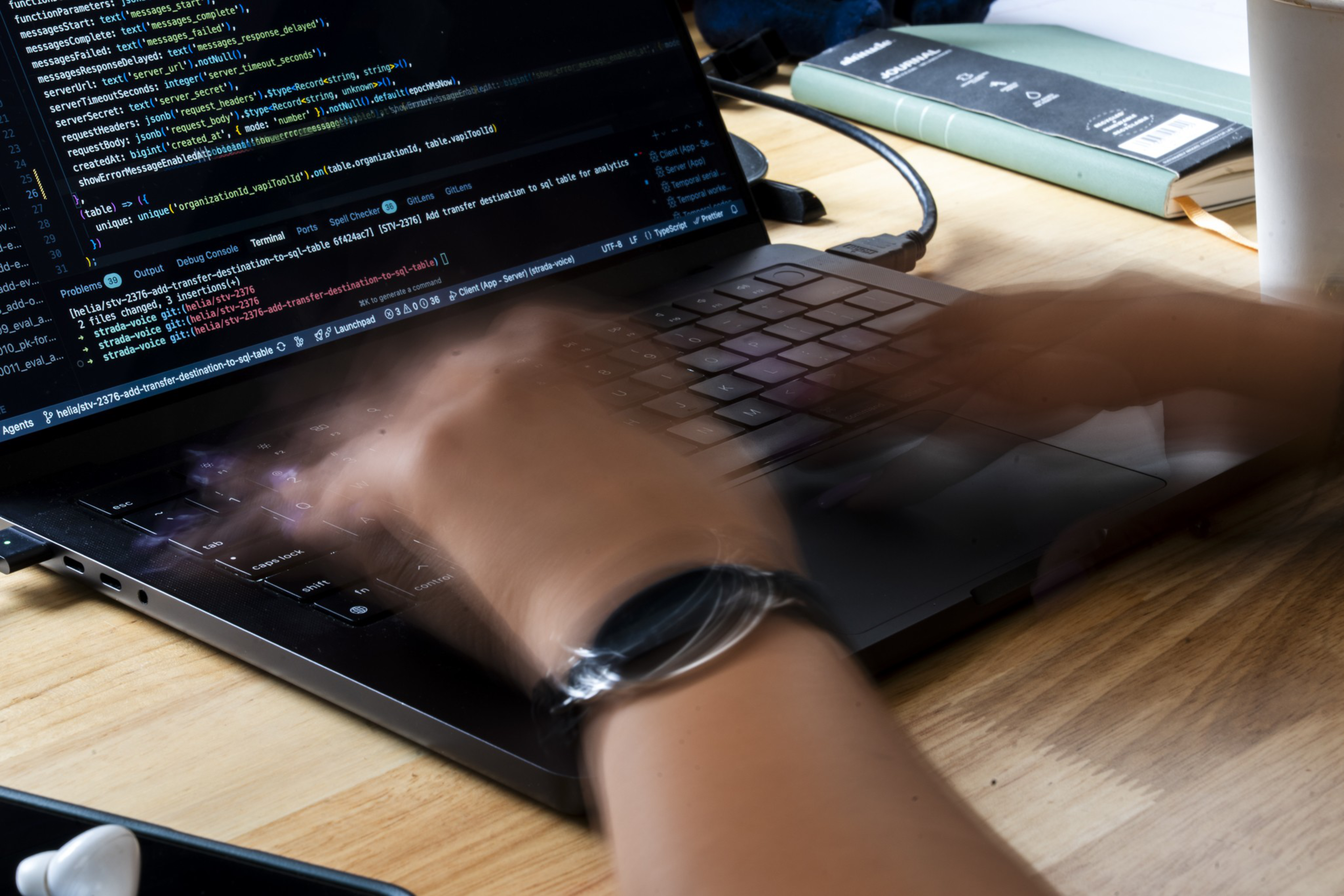 A person is typing quickly on a laptop displaying code, with a blurred motion effect on the hands, a book, and a cup are on the wooden desk.