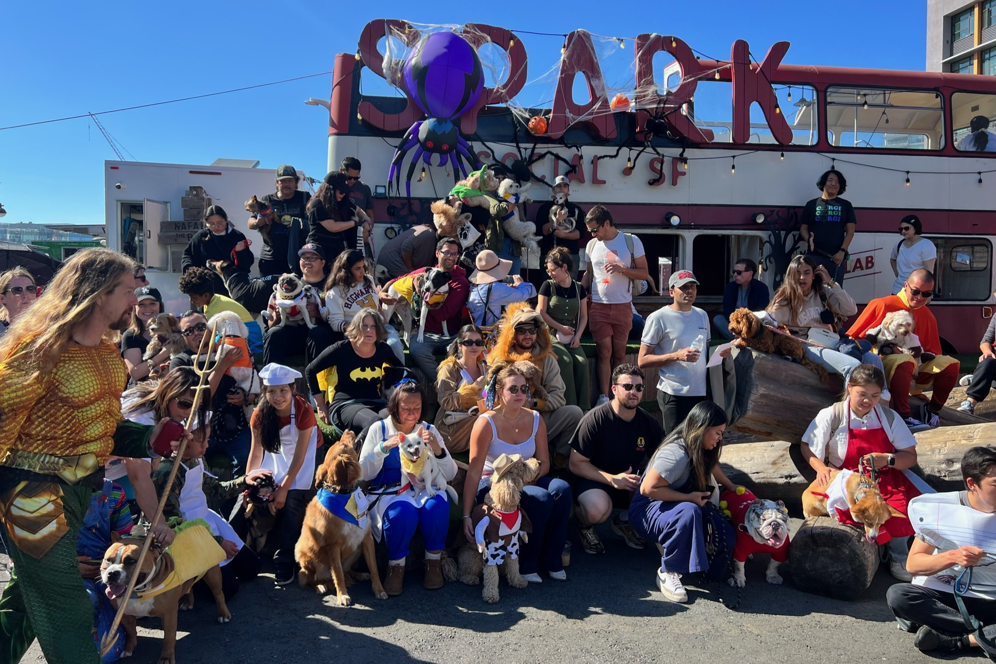 A large group of people and dogs, some in costumes, gather outdoors in front of a double-decker bus with Halloween decorations, including a large spider.