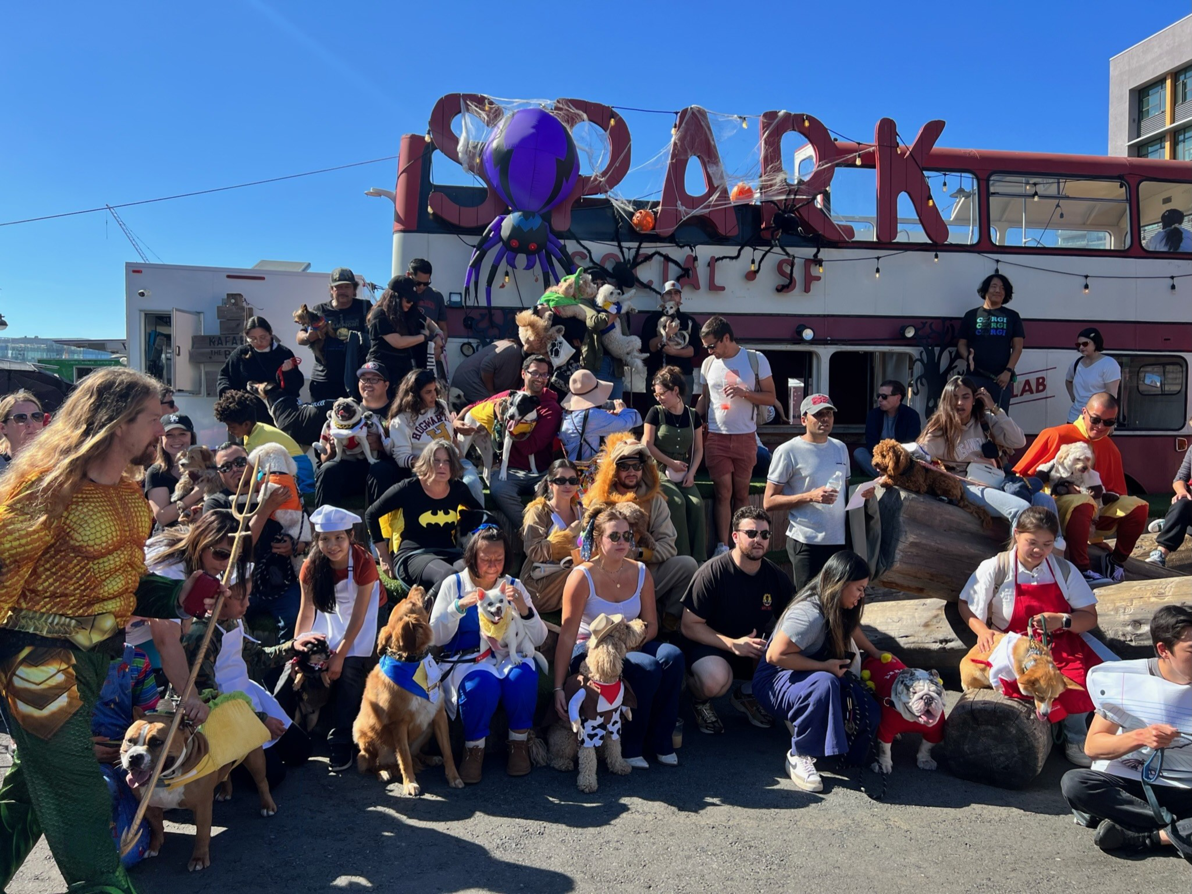 A large group of people and dogs, some in costumes, gather outdoors in front of a double-decker bus with Halloween decorations, including a large spider.