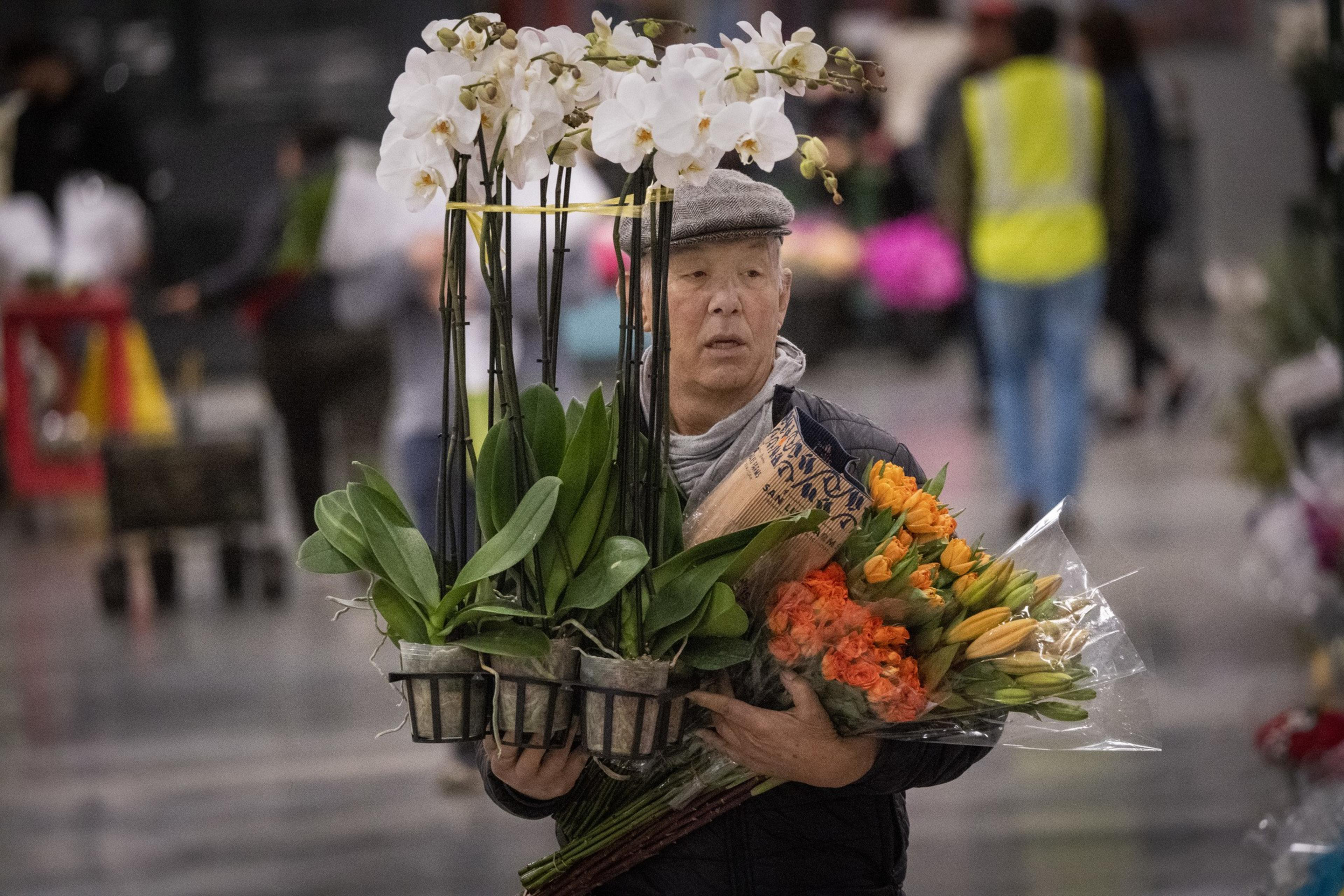 A man in a cap carries several potted orchids and bouquets of orange flowers in a busy market setting, with people and stalls blurred in the background.