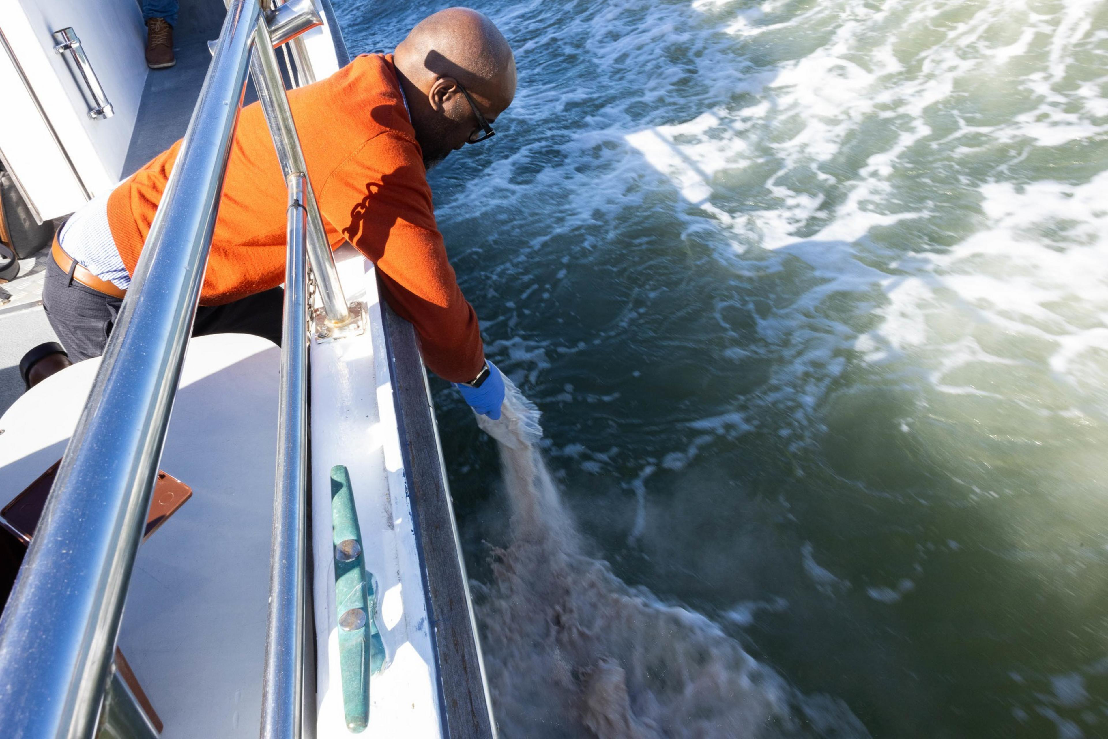 A man leans over the side of a boat, touching the water, with waves around. He drops ashes.