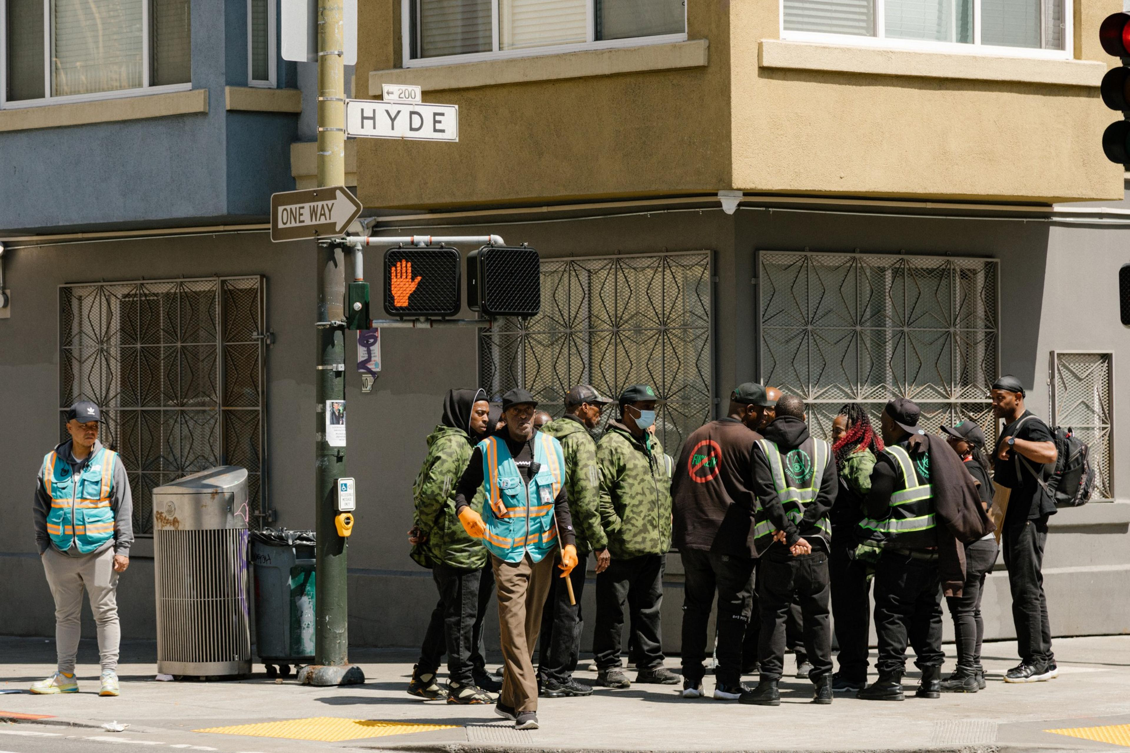 A group of people in vests and jackets stand by a street corner, with traffic lights indicating stop for pedestrians.