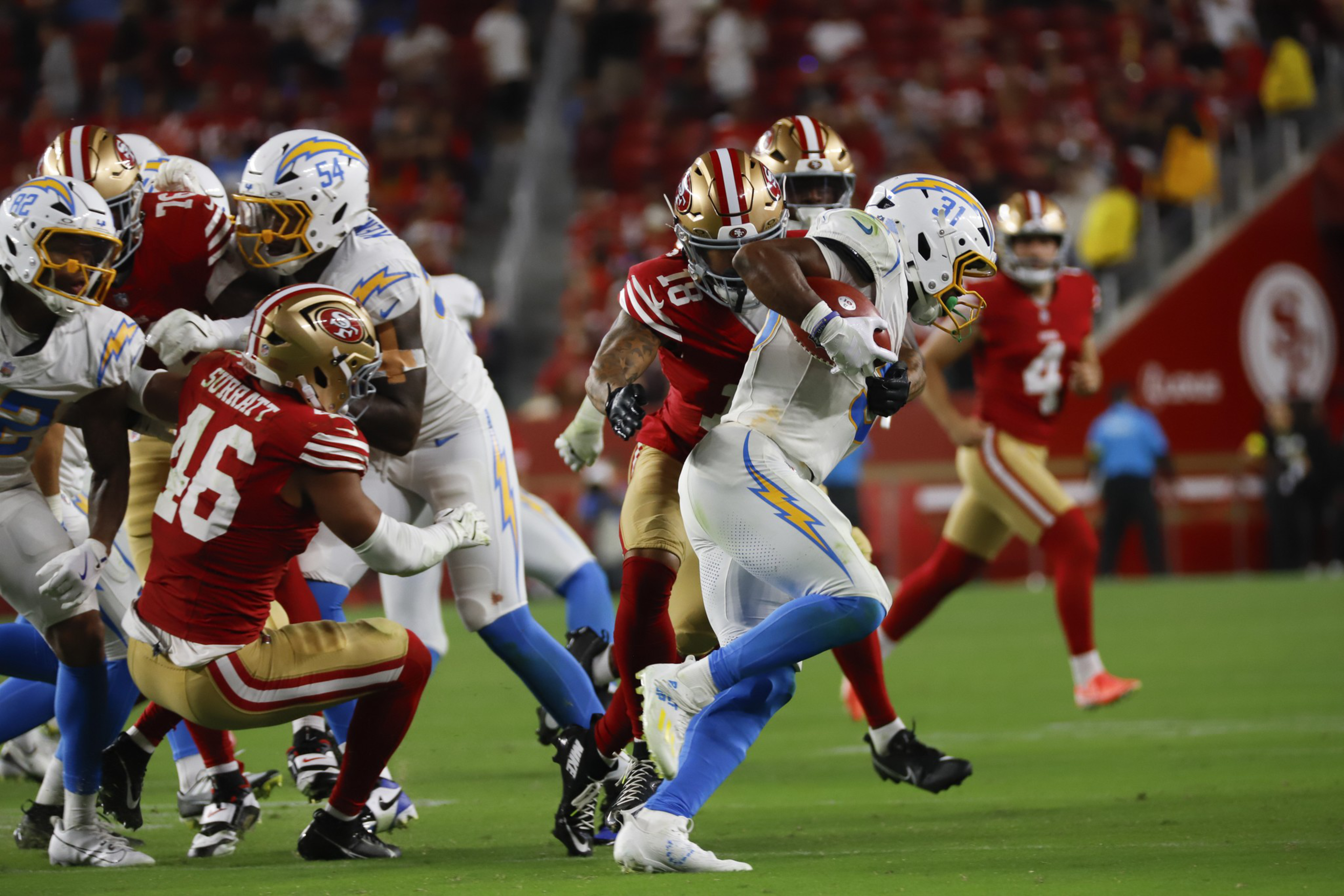 A football player in white and blue runs with the ball while a player in red and gold tries to tackle him during a game.