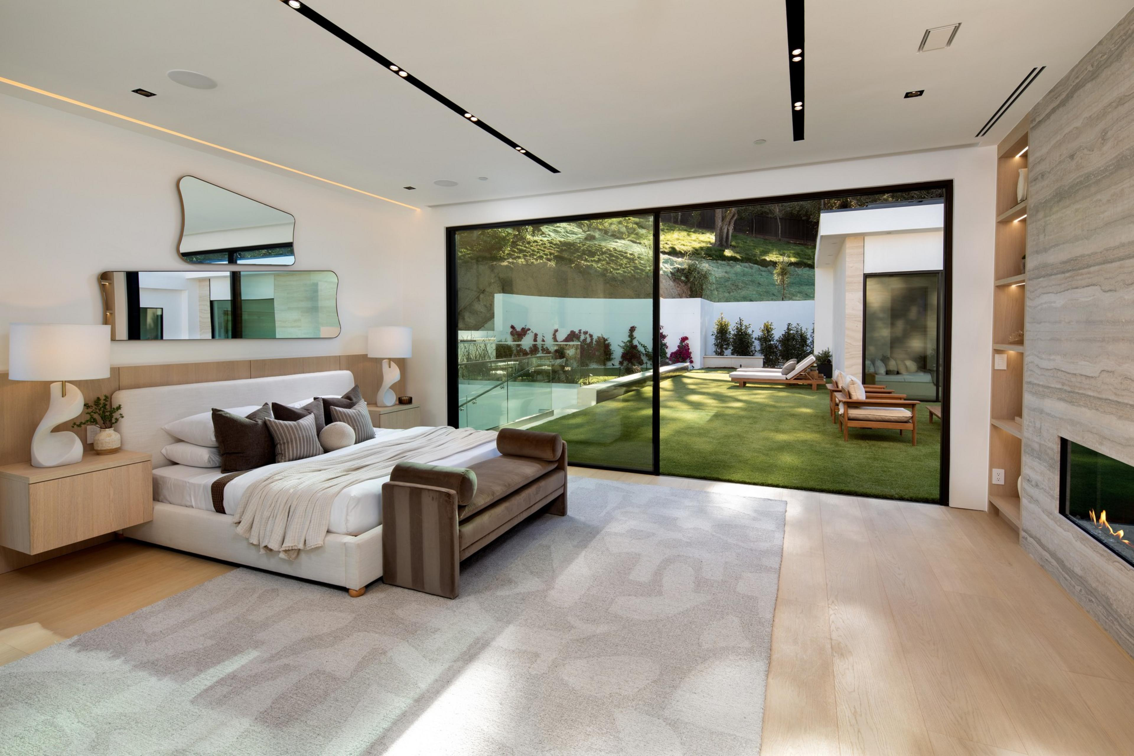 A modern bedroom features a large bed, leather bench, abstract mirrors, a fireplace, and sliding glass doors opening to a lawn with lounge chairs.