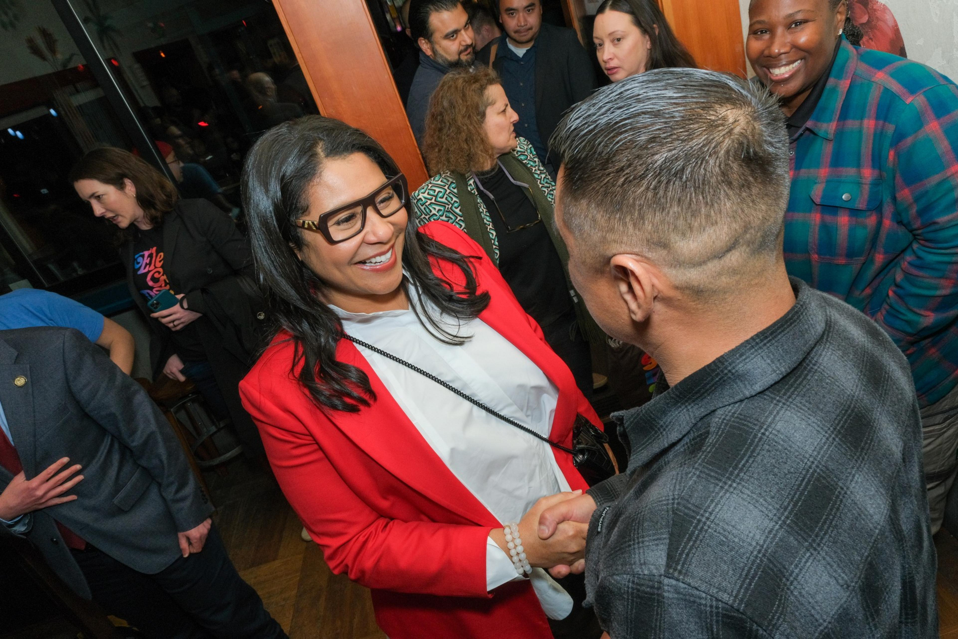 Mayor London Breed in a red blazer shakes hands with a man in a crowded room.