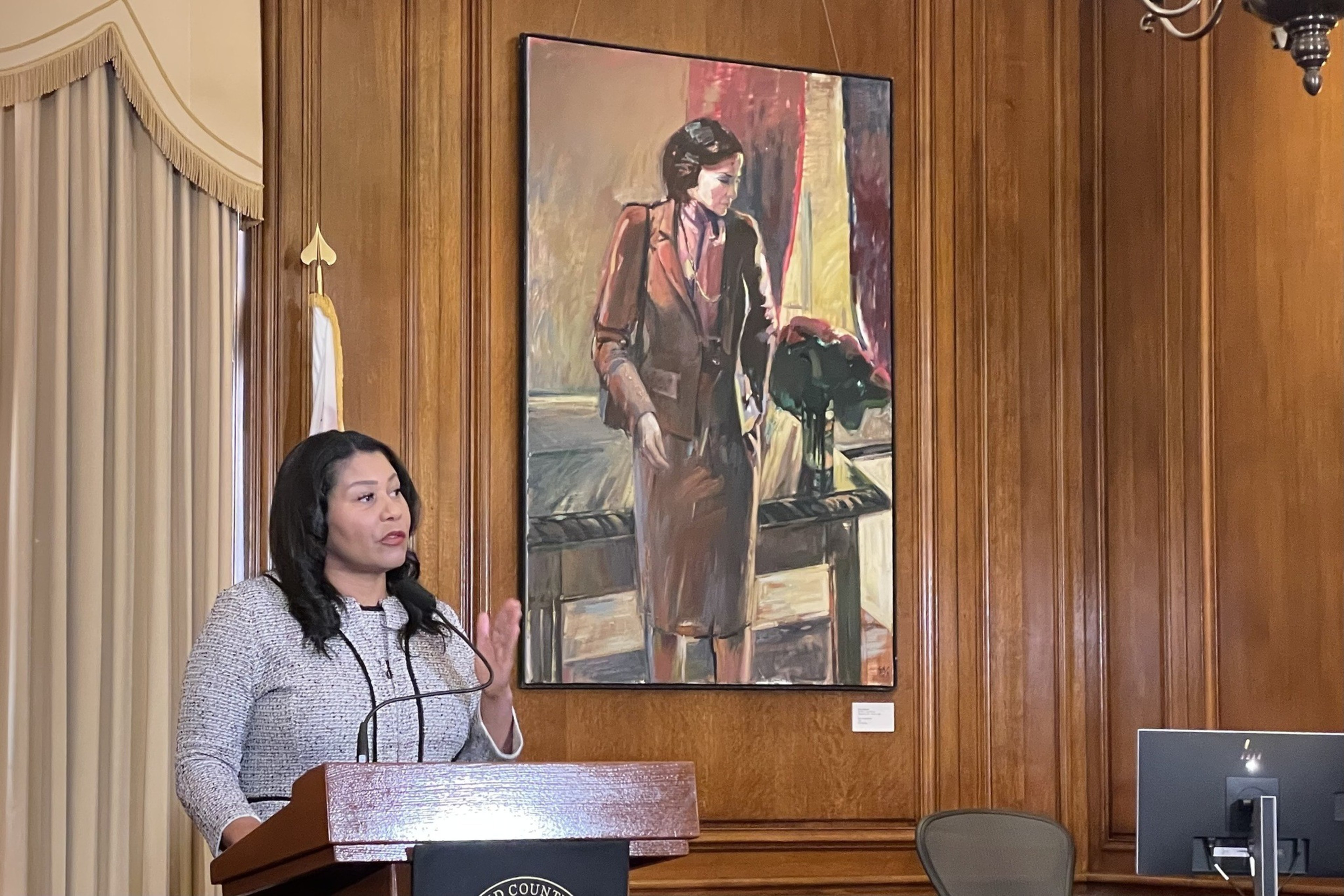 A woman speaks at a wooden podium in a wood-paneled room with a painting of a woman in a suit behind her and a computer on the side.