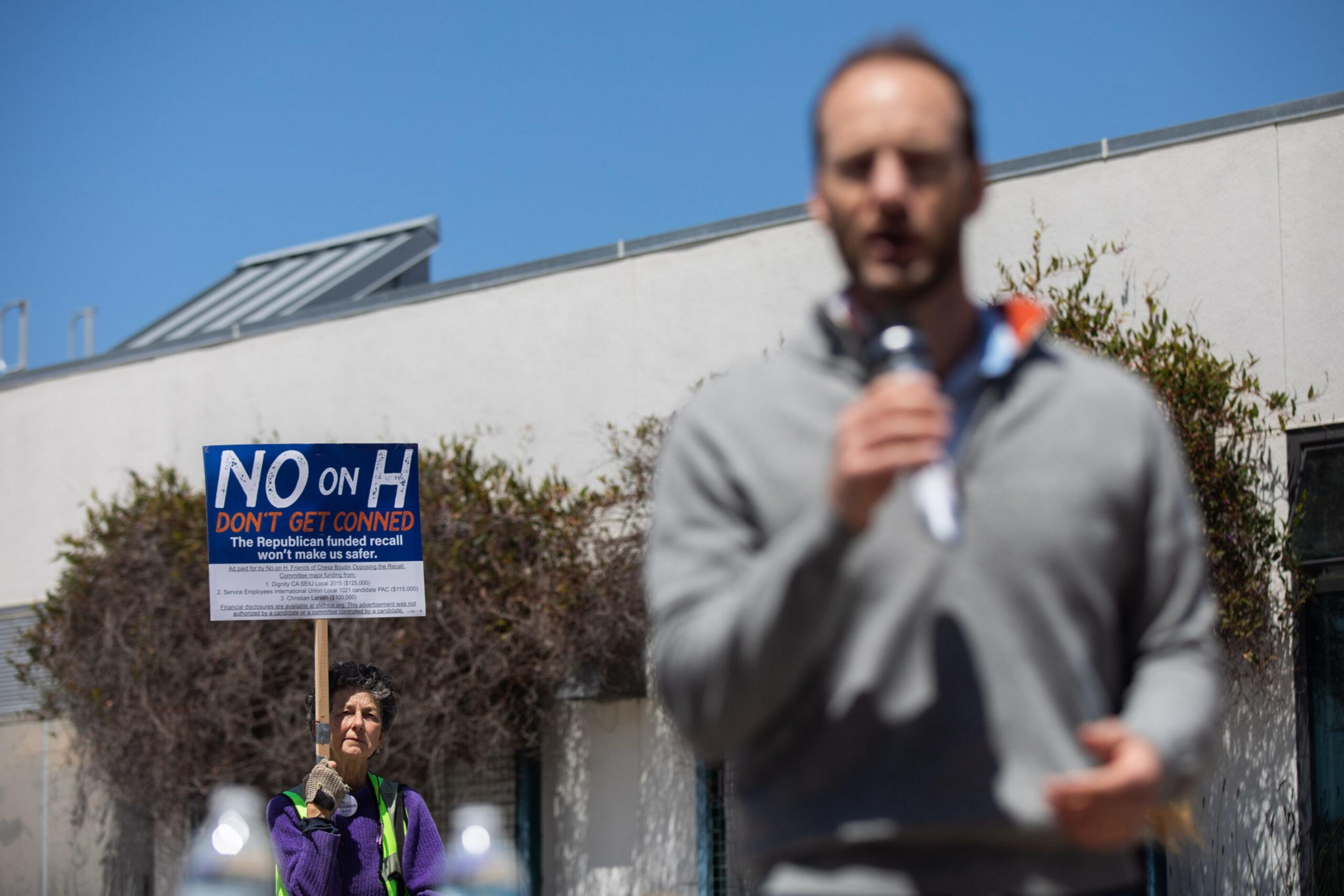 A person holds a sign that reads "NO on H, Don't Get Conned," while a blurred man speaks into a microphone in the foreground.