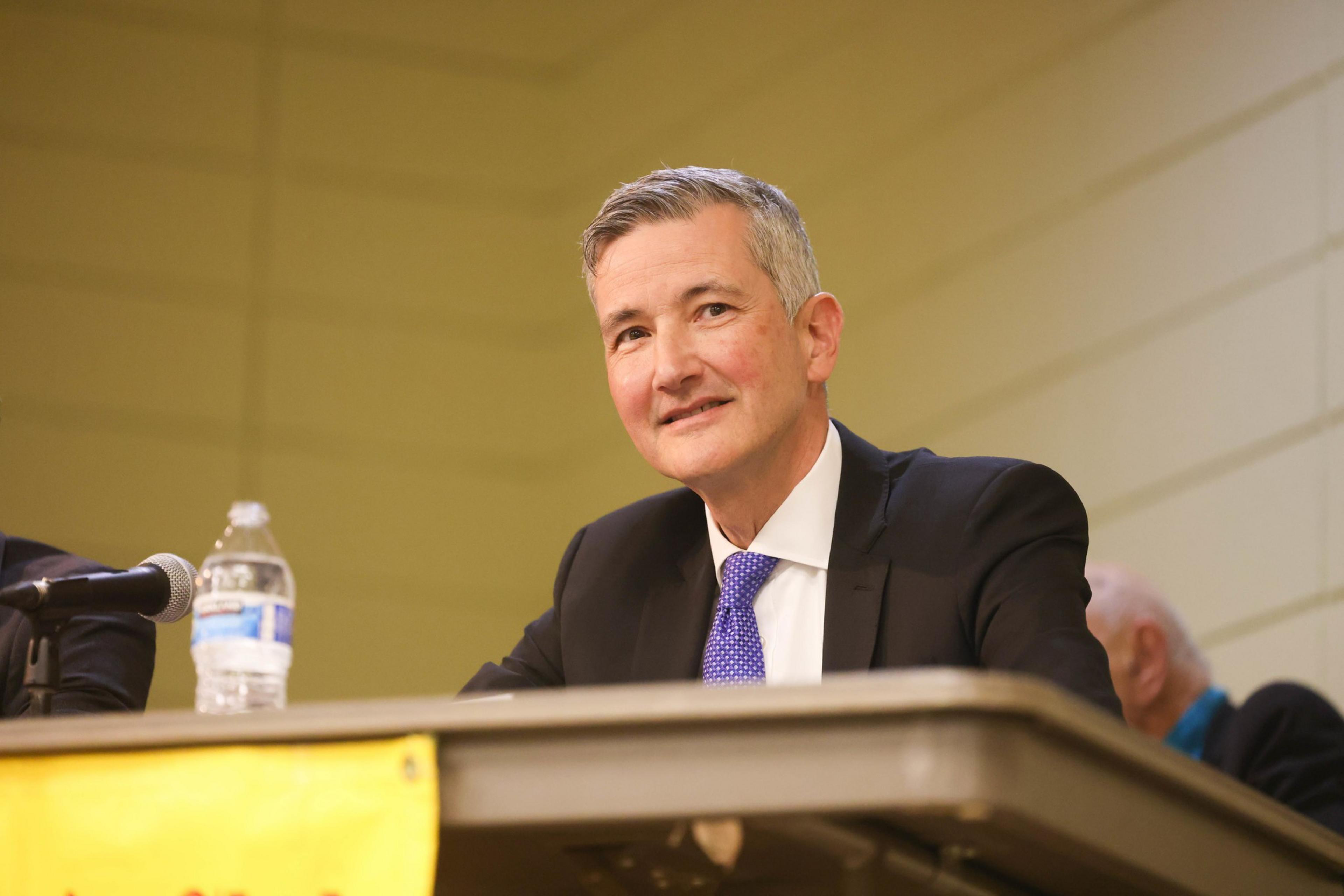 A man in a suit with a blue tie smiles while sitting at a table with a microphone and water bottle.