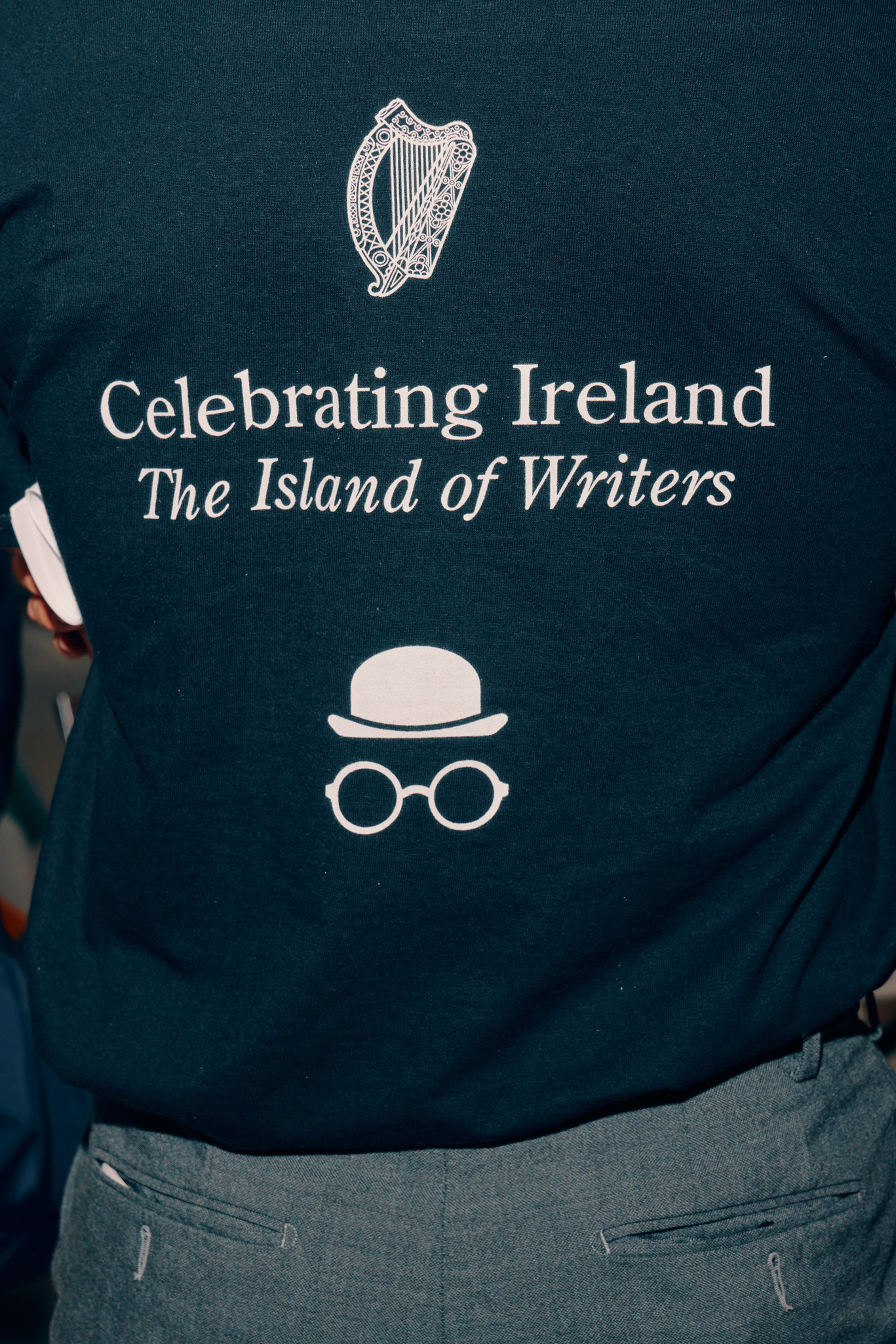 A black shirt with white text saying “Celebrating Ireland The Island of Writers,” featuring a harp above and a bowler hat with round glasses below.