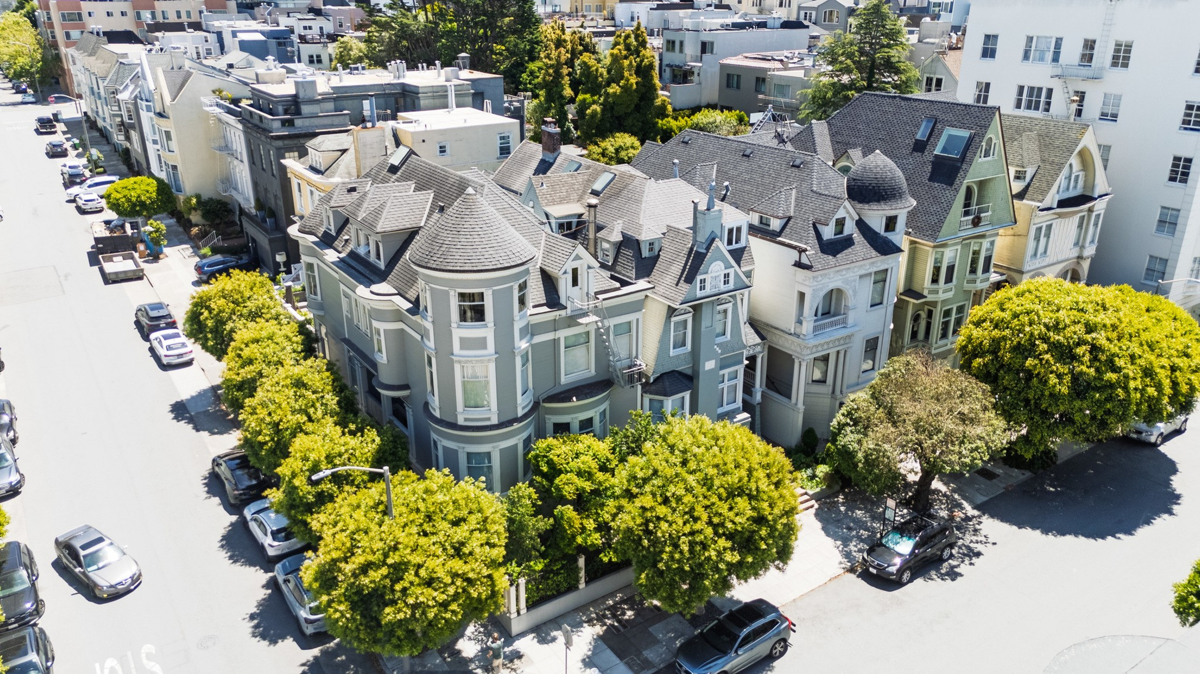 A street corner with multiple Victorian-style houses surrounded by lush green trees and parked cars on sunny day.