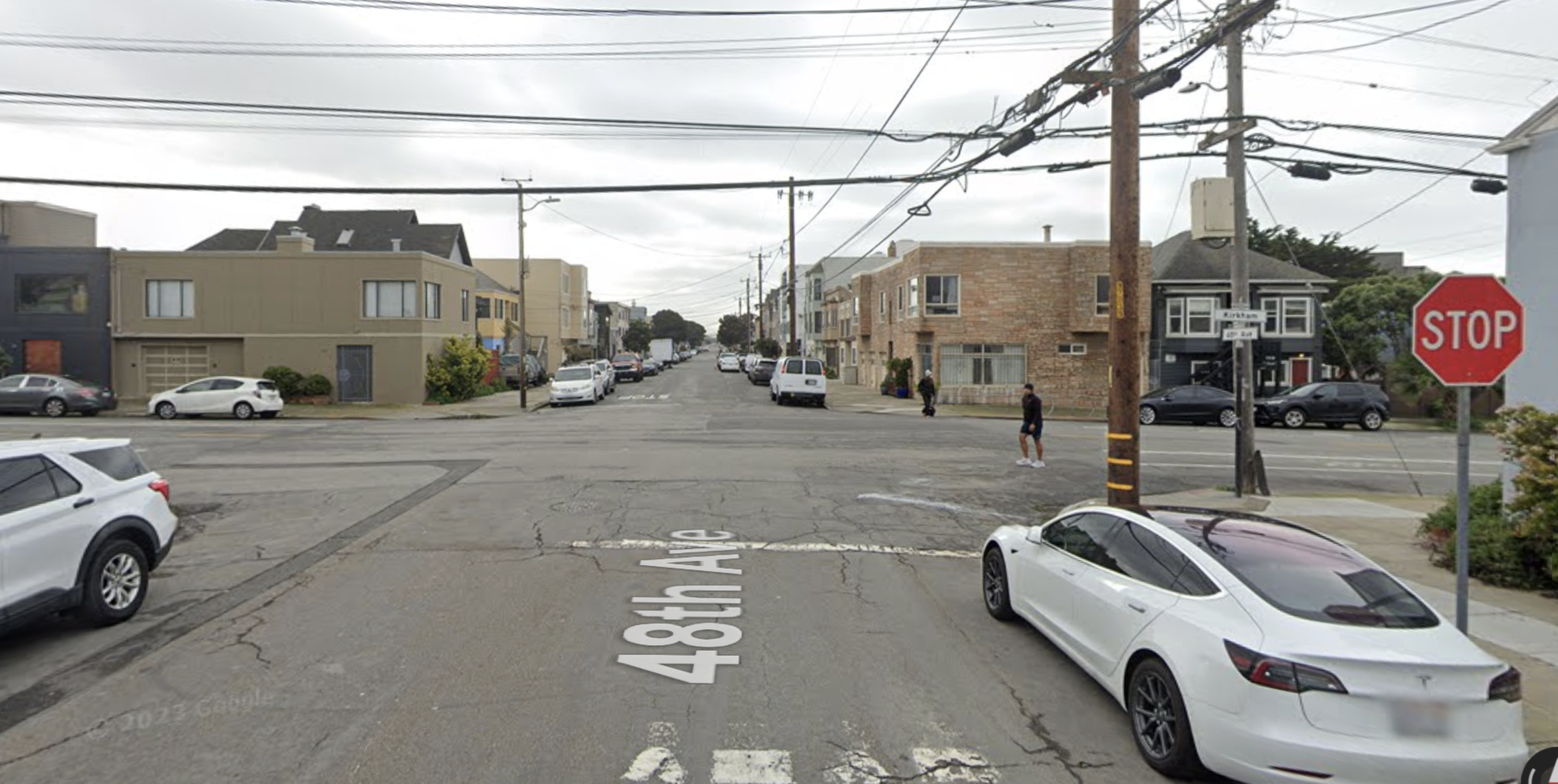 A street intersection with a visible "STOP" sign, white cars parked along the cracked road, and residential buildings lining both sides.