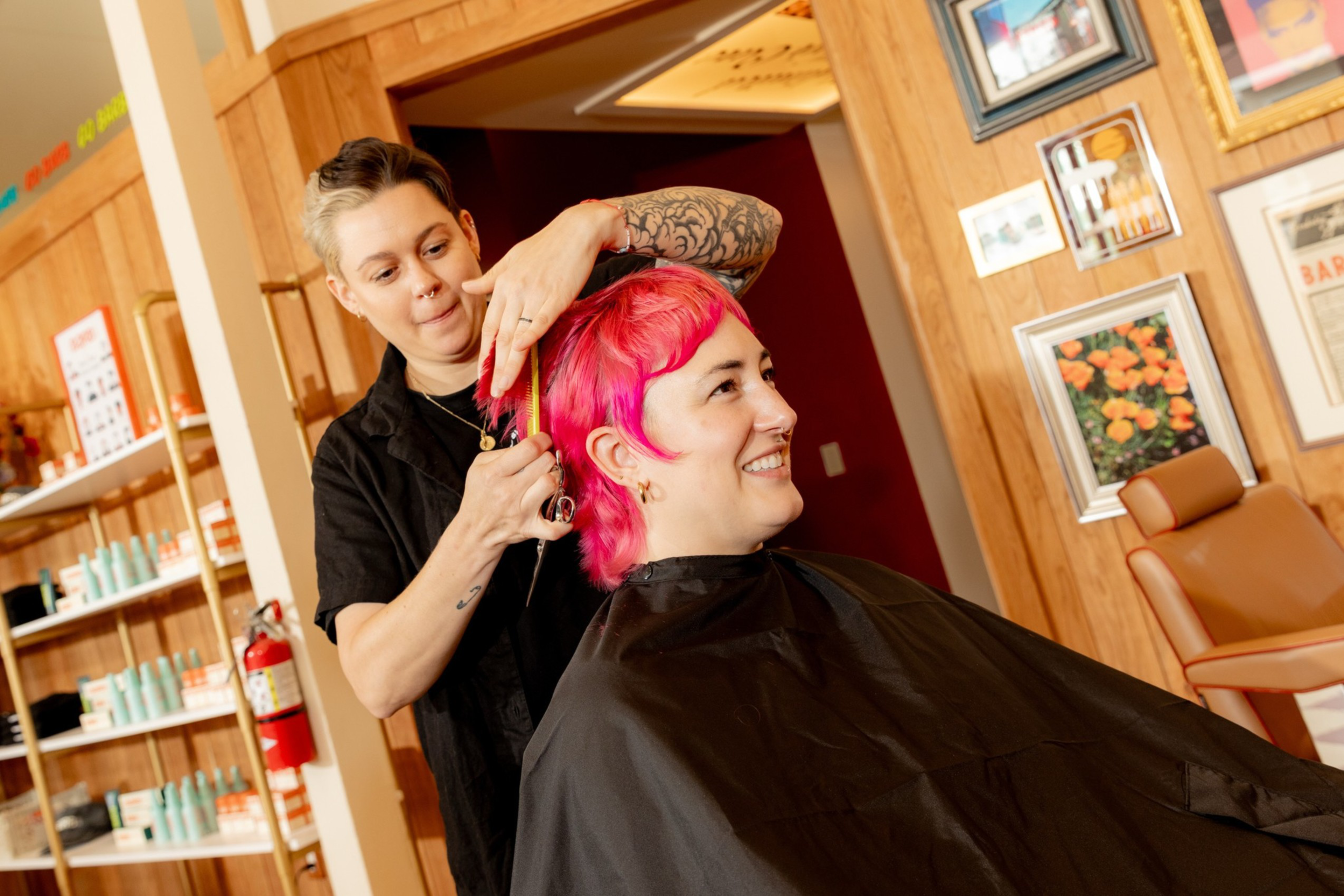 A tattooed hairstylist trims the bright pink hair of a smiling client wearing a black cape in a warmly decorated salon.