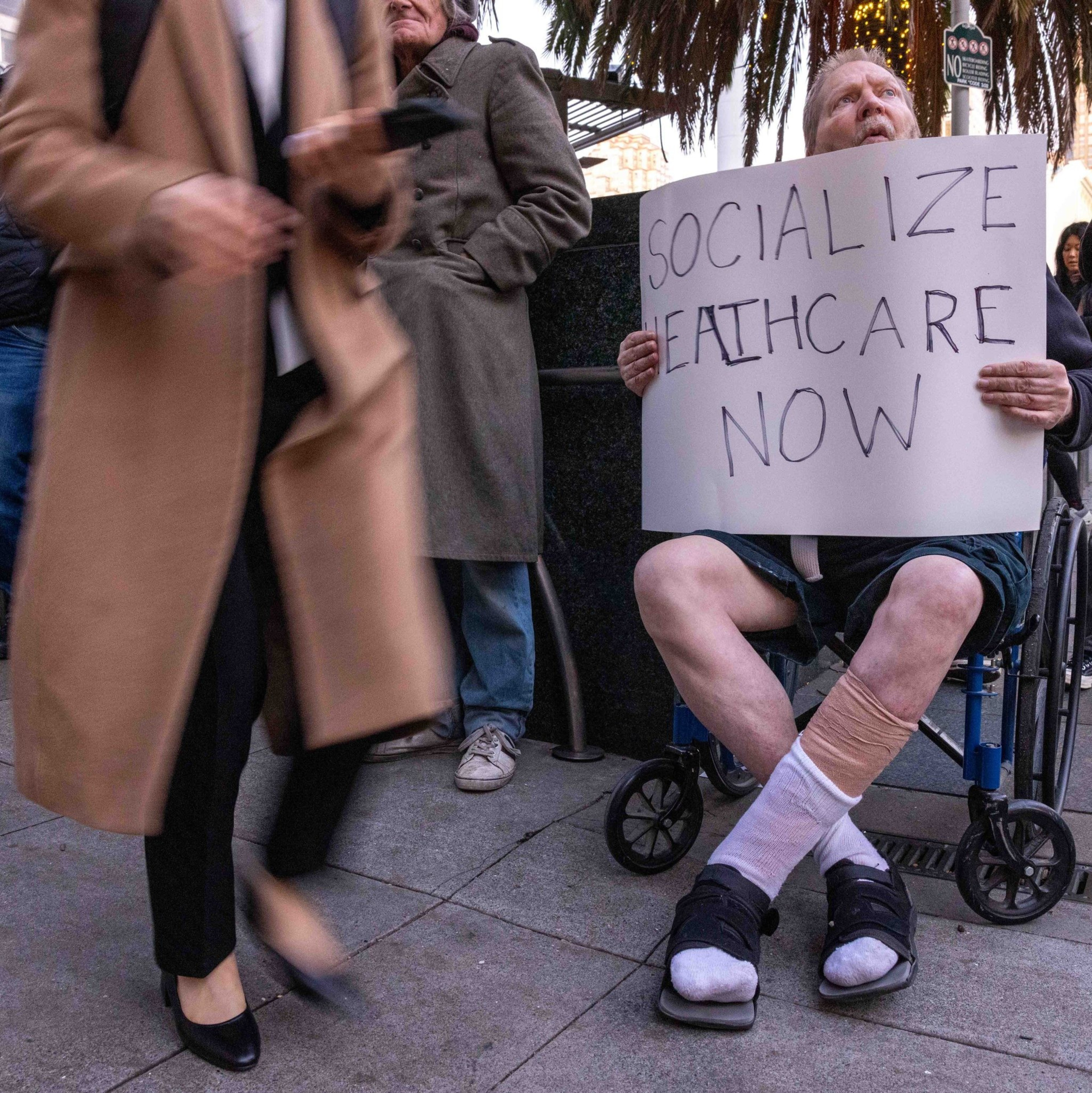 A person in a wheelchair holds a sign saying &quot;Socialize Healthcare Now,&quot; surrounded by people on a city street. Another sign reads &quot;Stop [?]&quot; in the background.