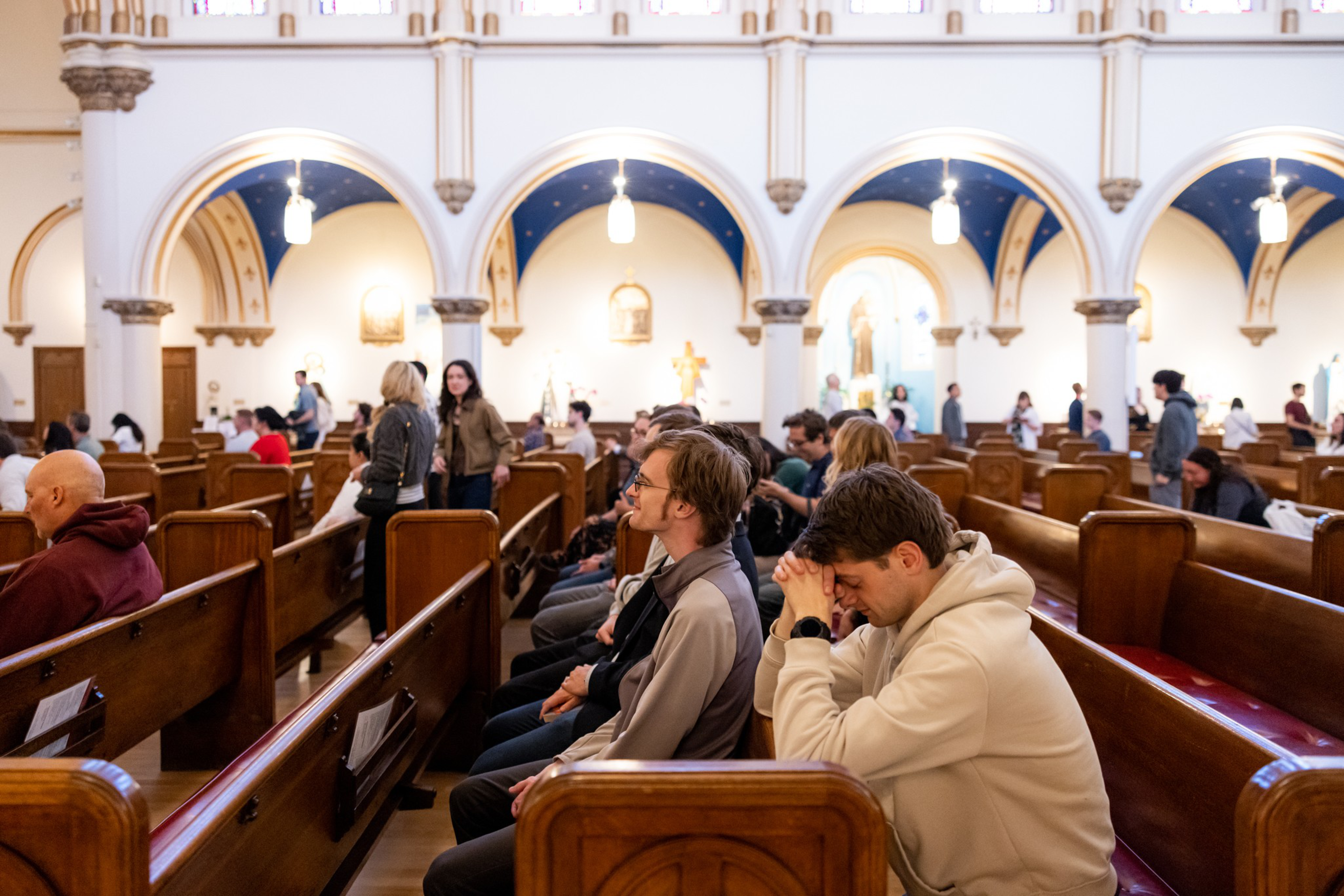 People seated on wooden pews inside a church, some praying, with arched ceilings and religious statues in the background.