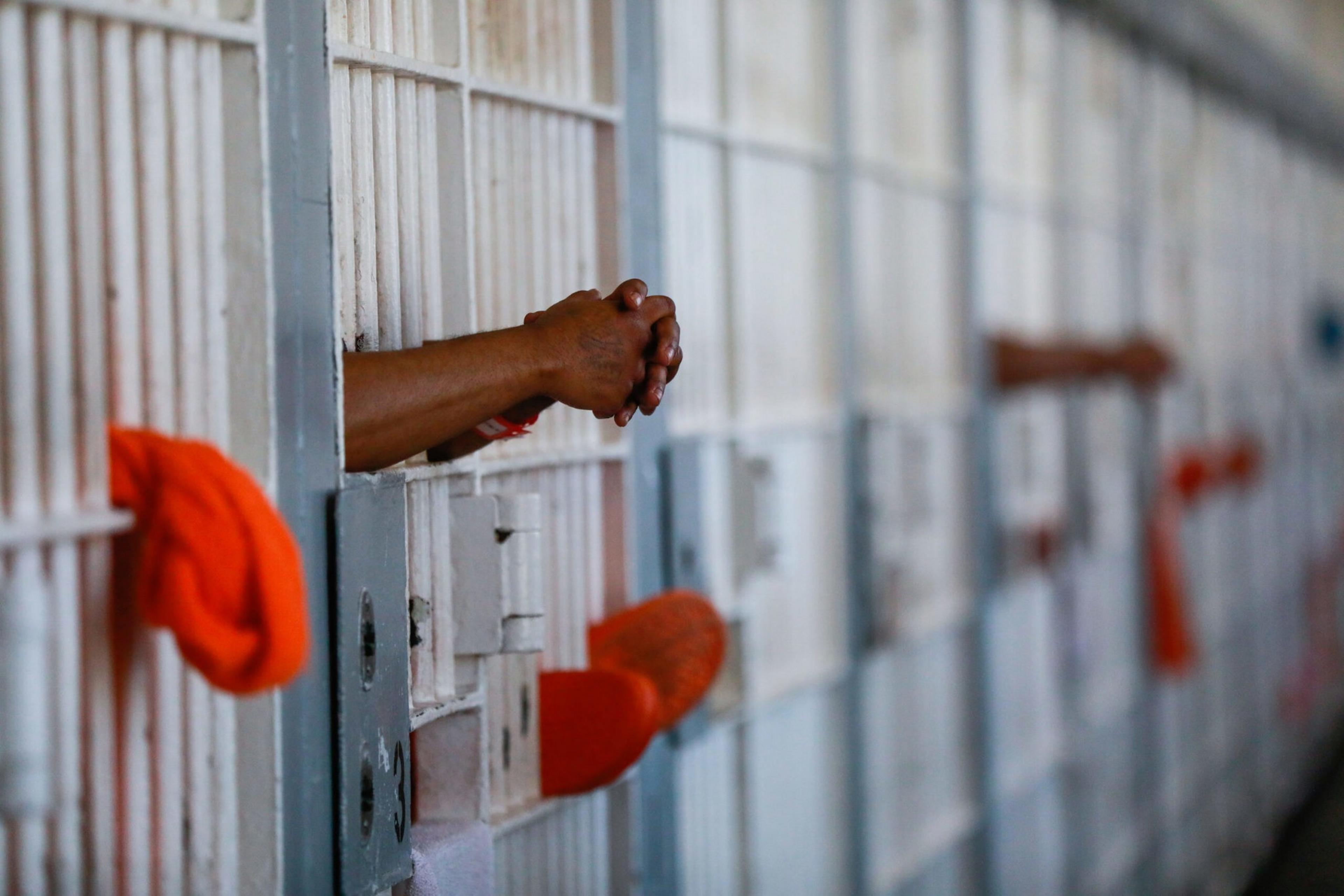 Hands and feet of prisoners extend through the bars of multiple jail cells, with several wearing orange clothing inside the cells.
