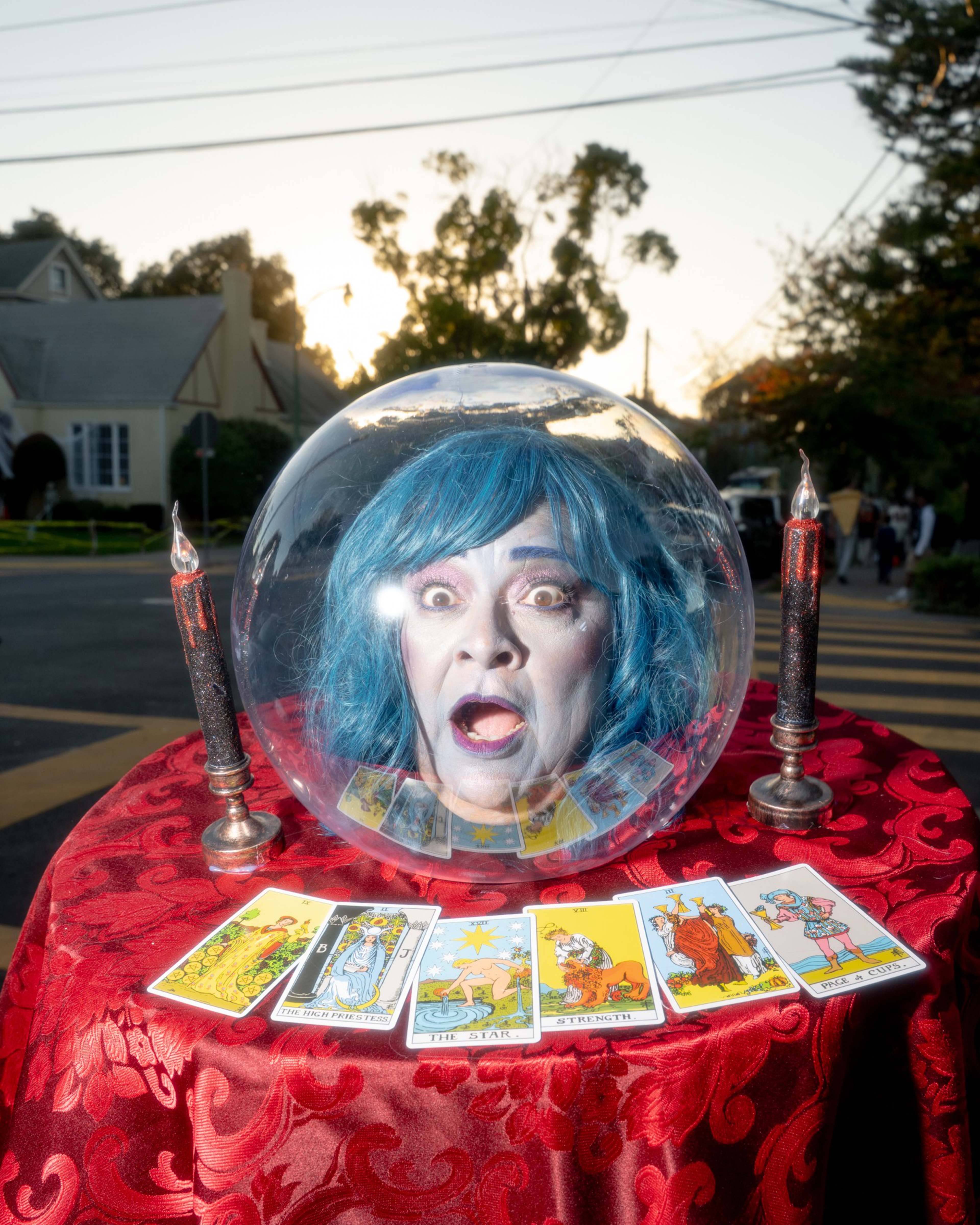 A blue-haired head with a surprised expression is inside a clear orb on a red table with tarot cards and two black candles.