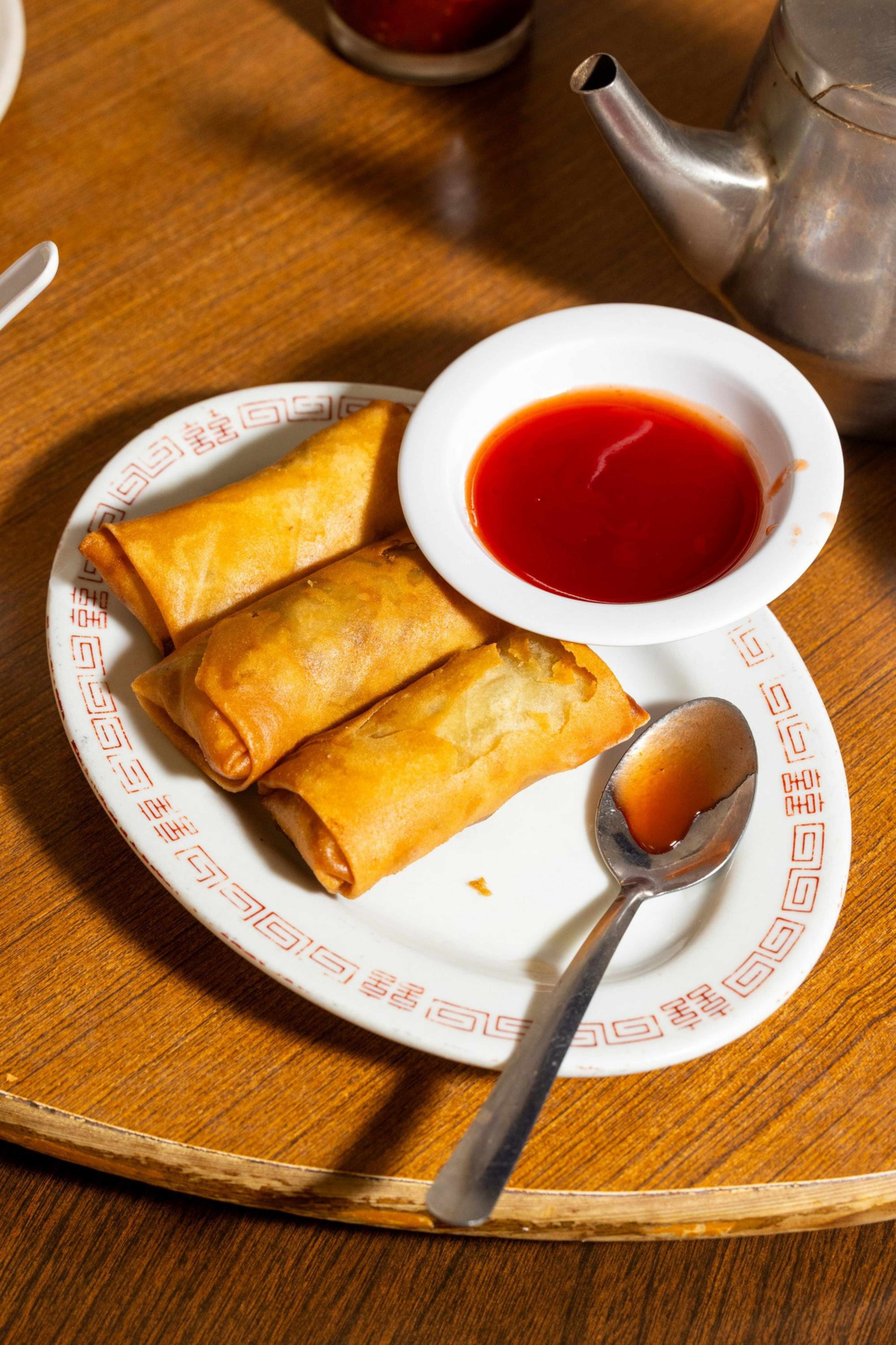 Three golden-brown spring rolls on a white oval plate with red sauce in a small bowl and a spoon with some sauce on a wooden table.