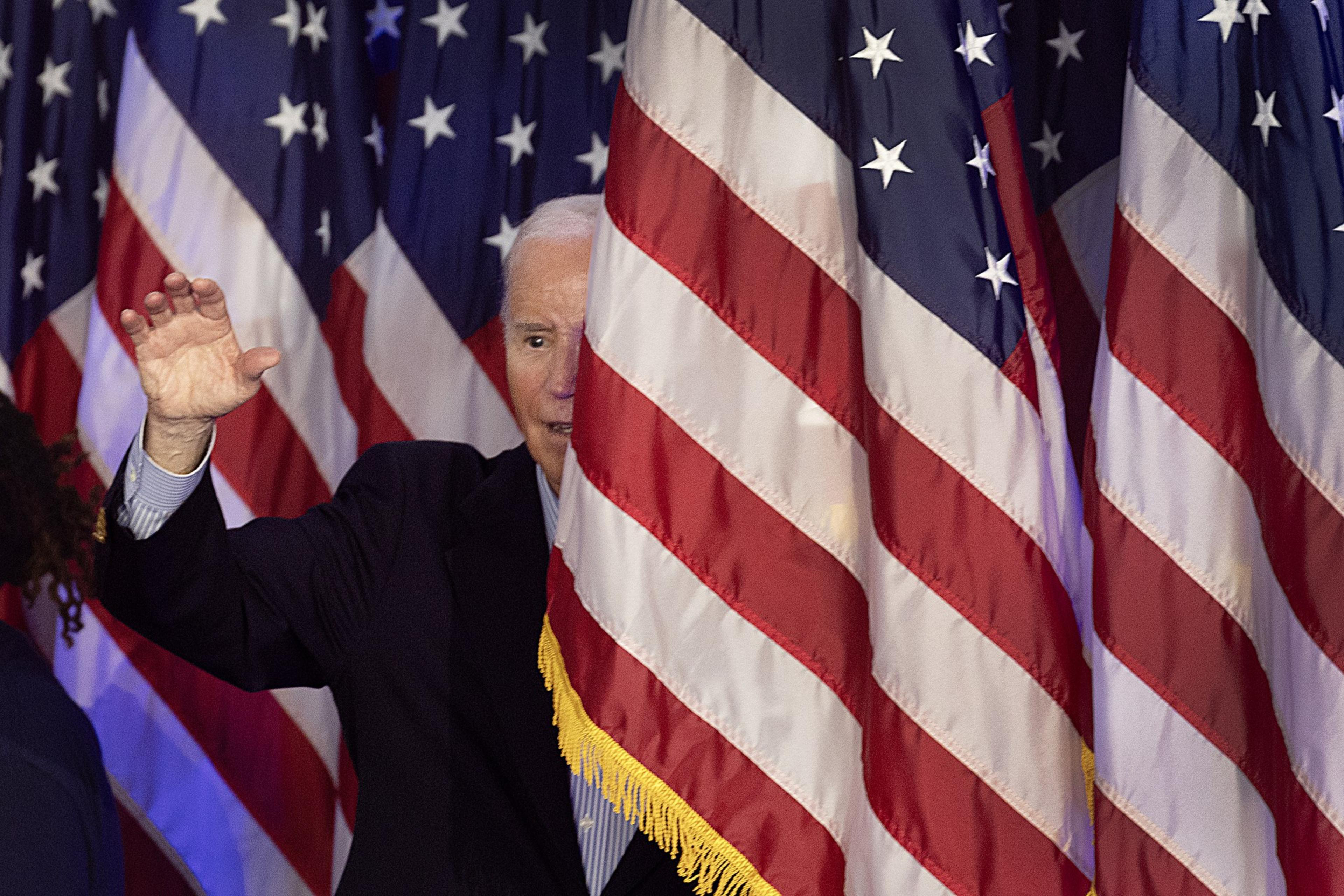 A person dressed in a dark suit is partially obscured by several American flags, with only part of their face and a raised hand visible.