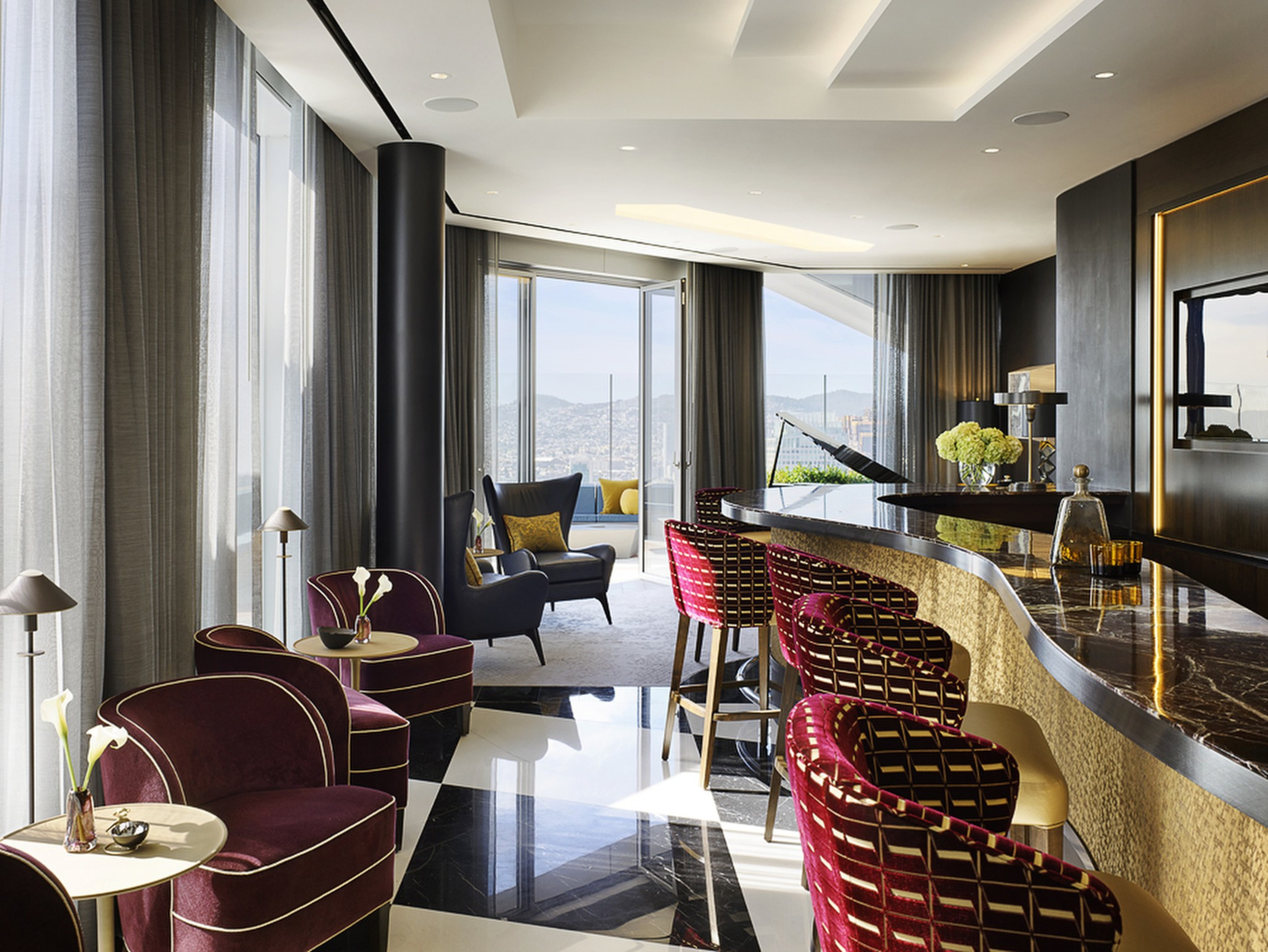 A modern lounge features a curved marble bar with red cushioned stools, velvet armchairs, small tables with flowers, sheer curtains, and floor-to-ceiling windows.