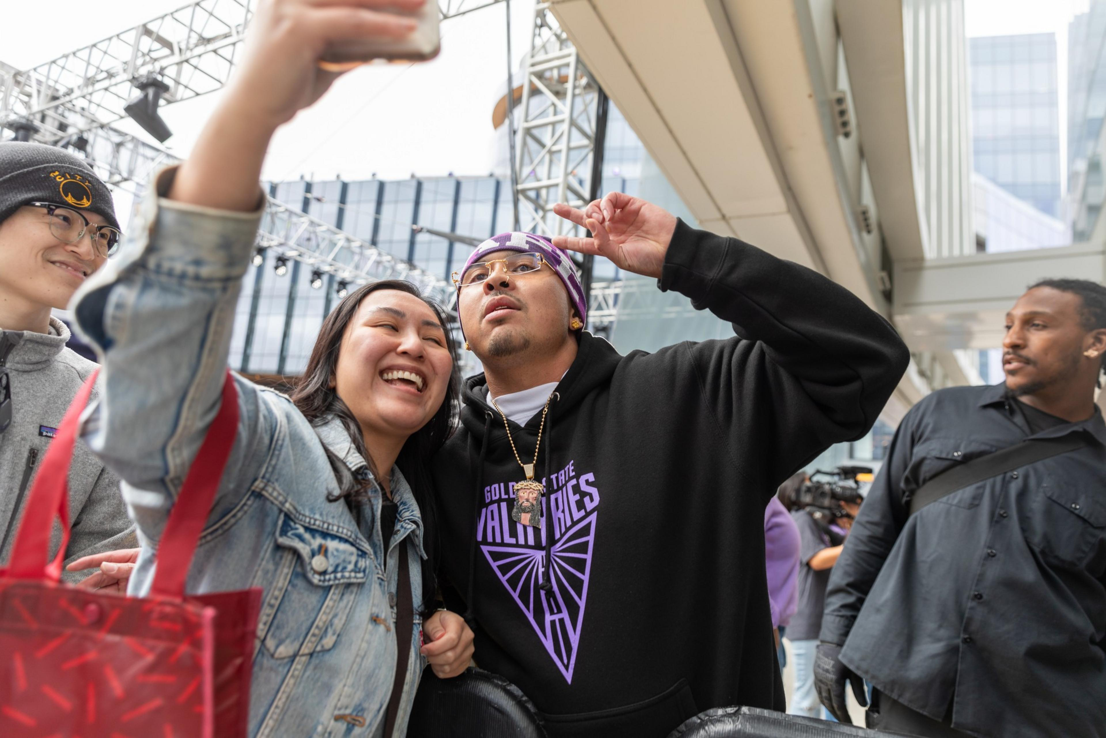 Three people are joyfully taking a selfie outdoors; one man makes a peace sign, the woman smiles widely. A bystander observes, set against an urban backdrop.
