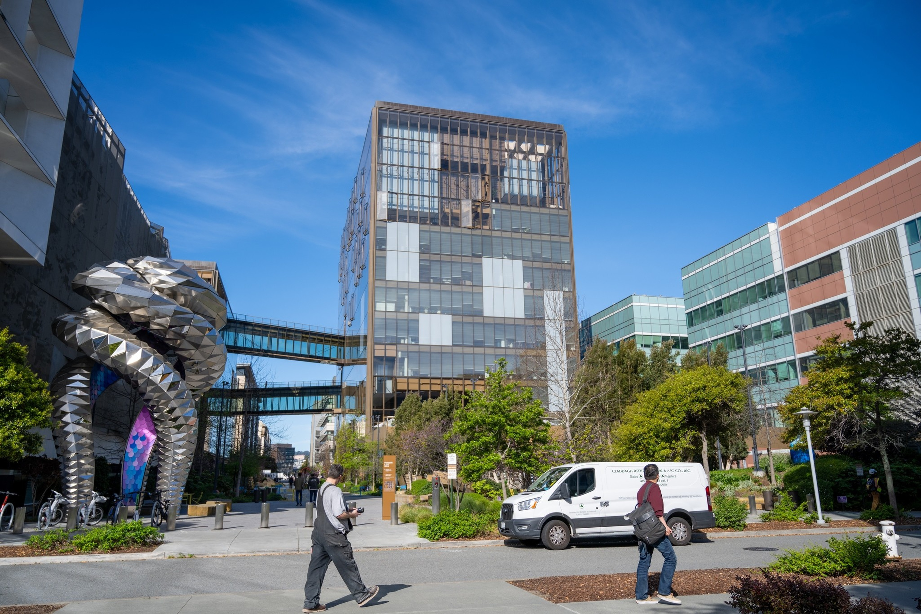 Two people walk near modern office buildings with glass facades on a sunny day, alongside a distinctive metallic sculpture and a white van.