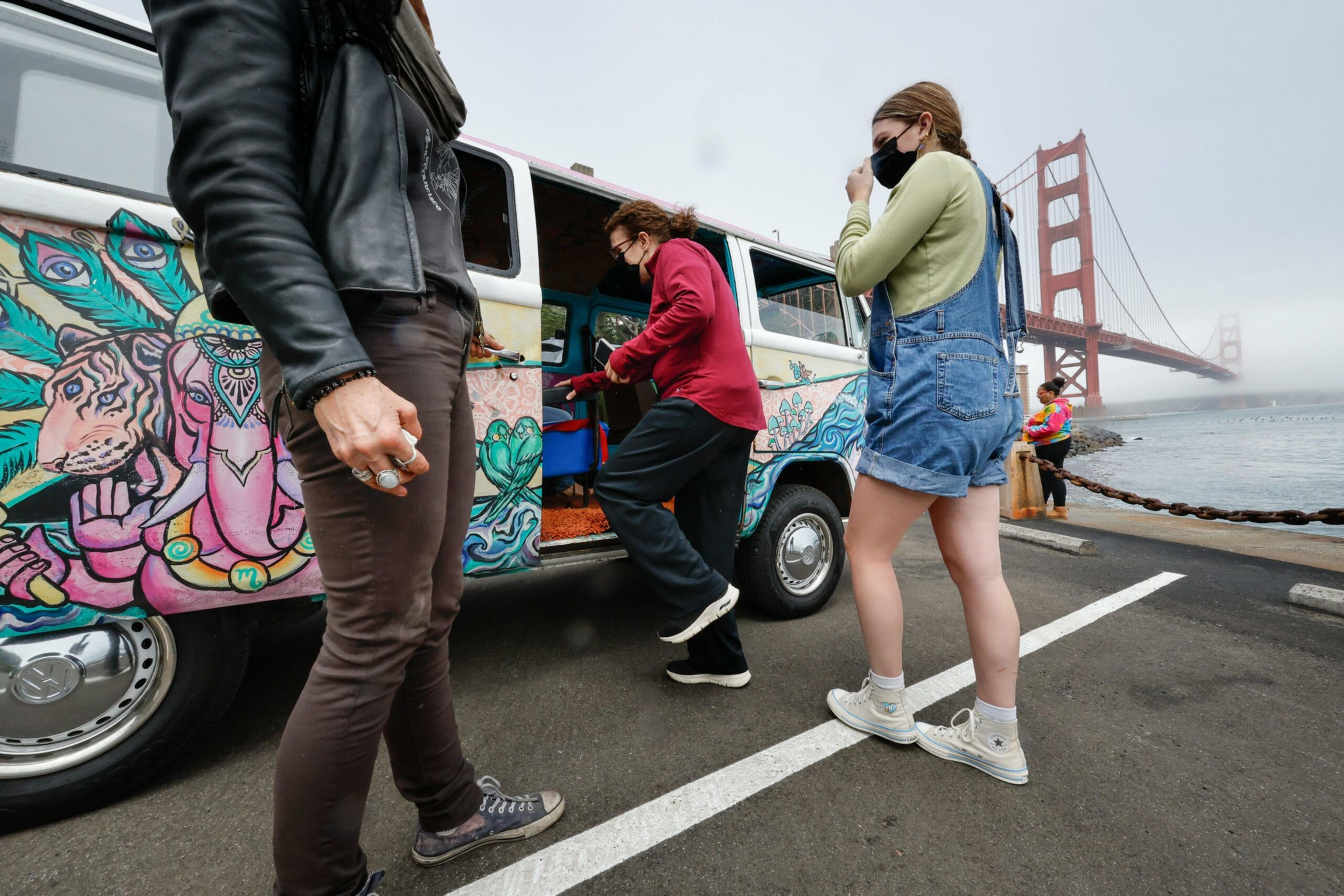 Three people stand near a colorfully painted van by the water with the Golden Gate Bridge in the background. One person is entering the van, and two are standing outside.