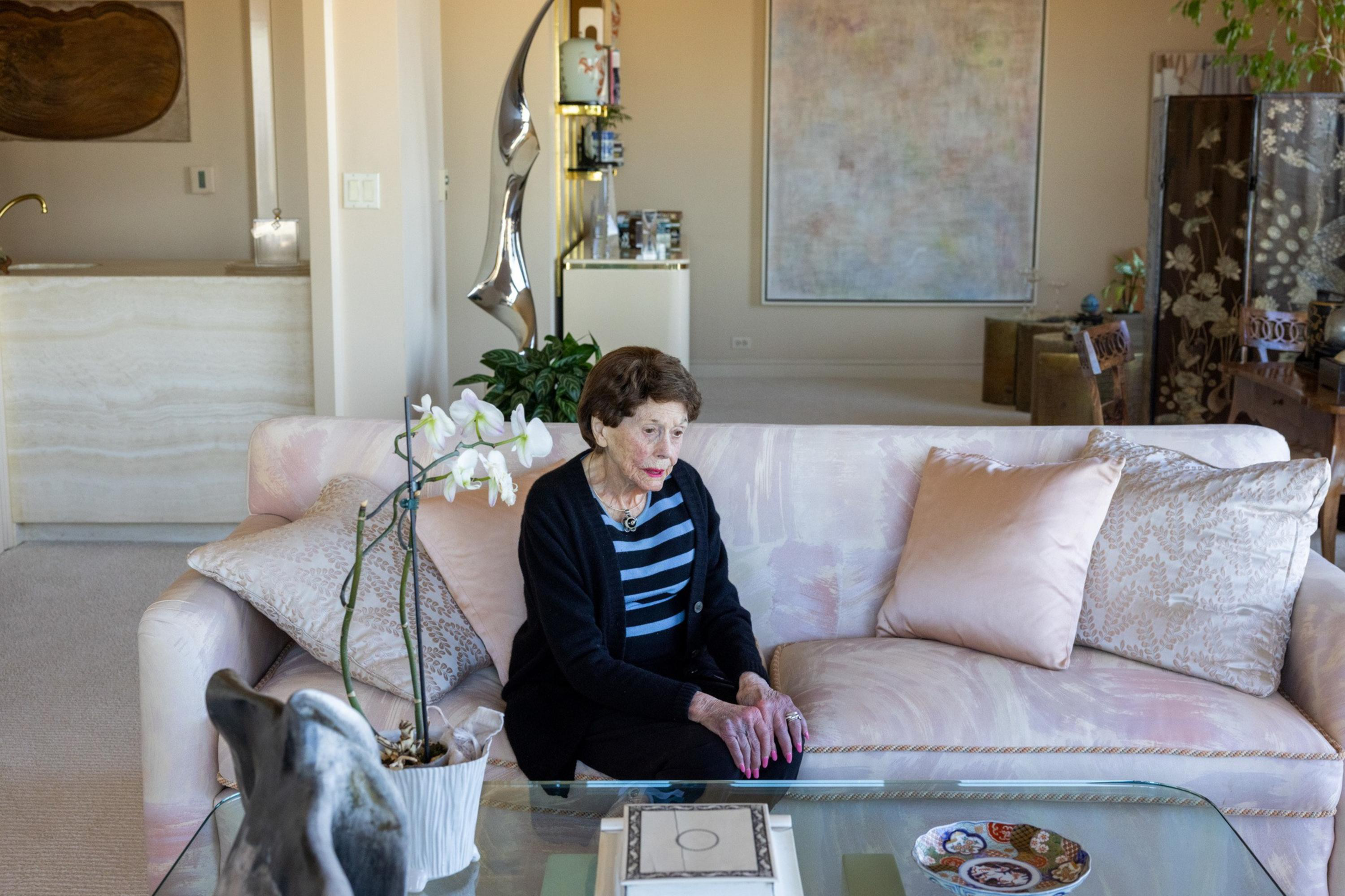 An elderly woman with short brown hair sits on a pale pink sofa surrounded by light-colored cushions in a well-lit living room.