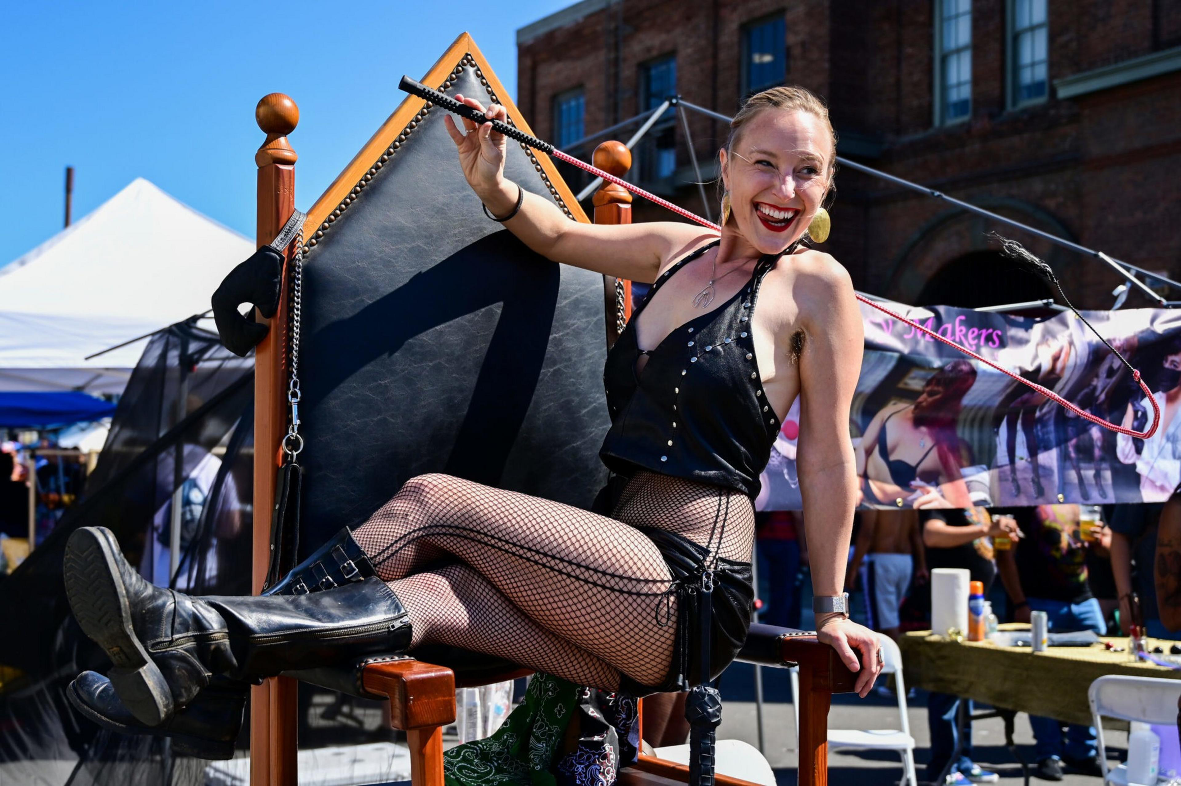 A smiling woman in a black leather outfit and fishnet stockings lounges confidently on a tall wooden throne outdoors, holding a riding crop.