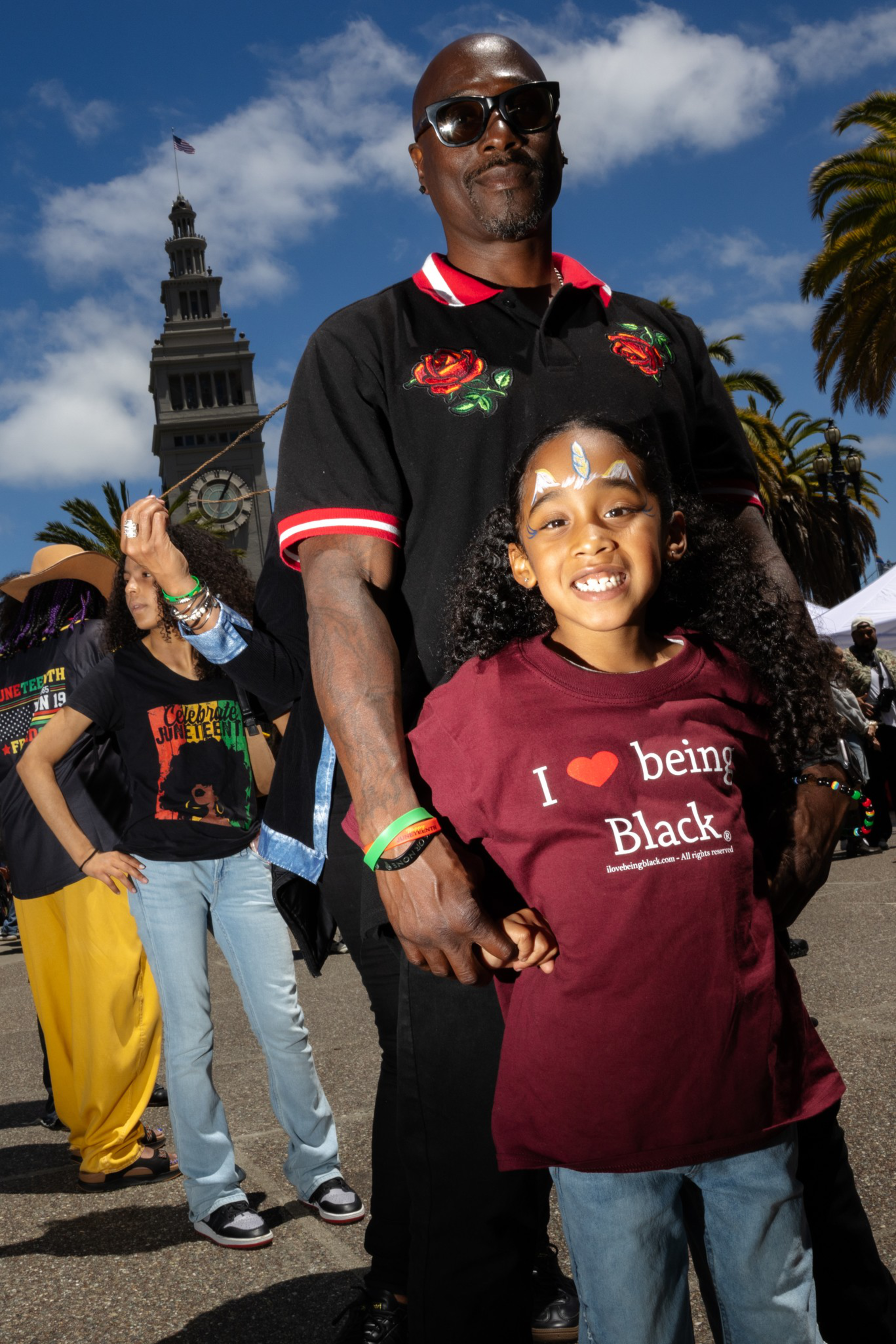 A smiling man in sunglasses stands behind a girl with face paint wearing a maroon “I love being Black” shirt, with others and a clock tower in the background.