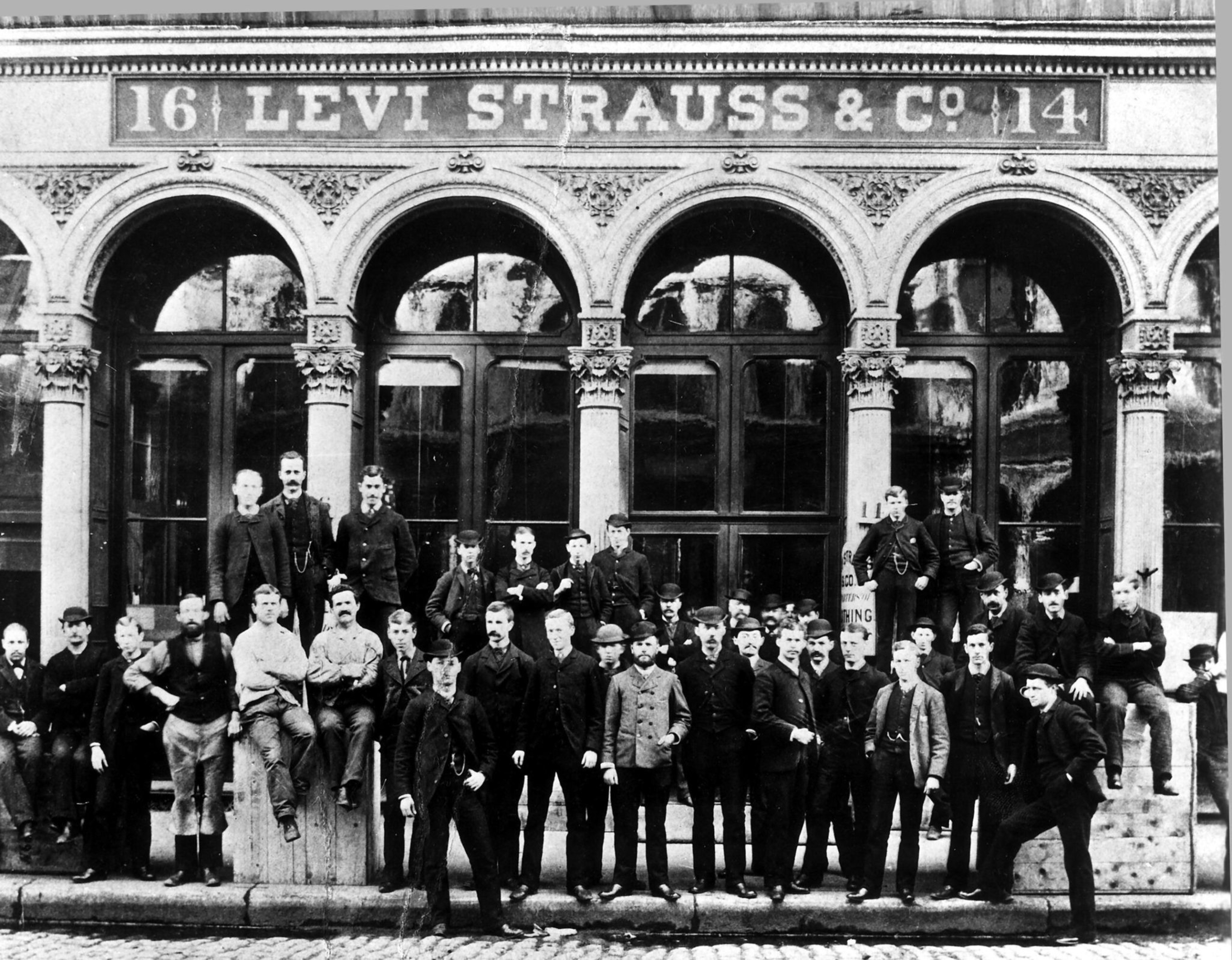 A group of about 40 men in late 19th-century clothing pose in front of a building with arched windows and a sign that reads "Levi Strauss & Co."