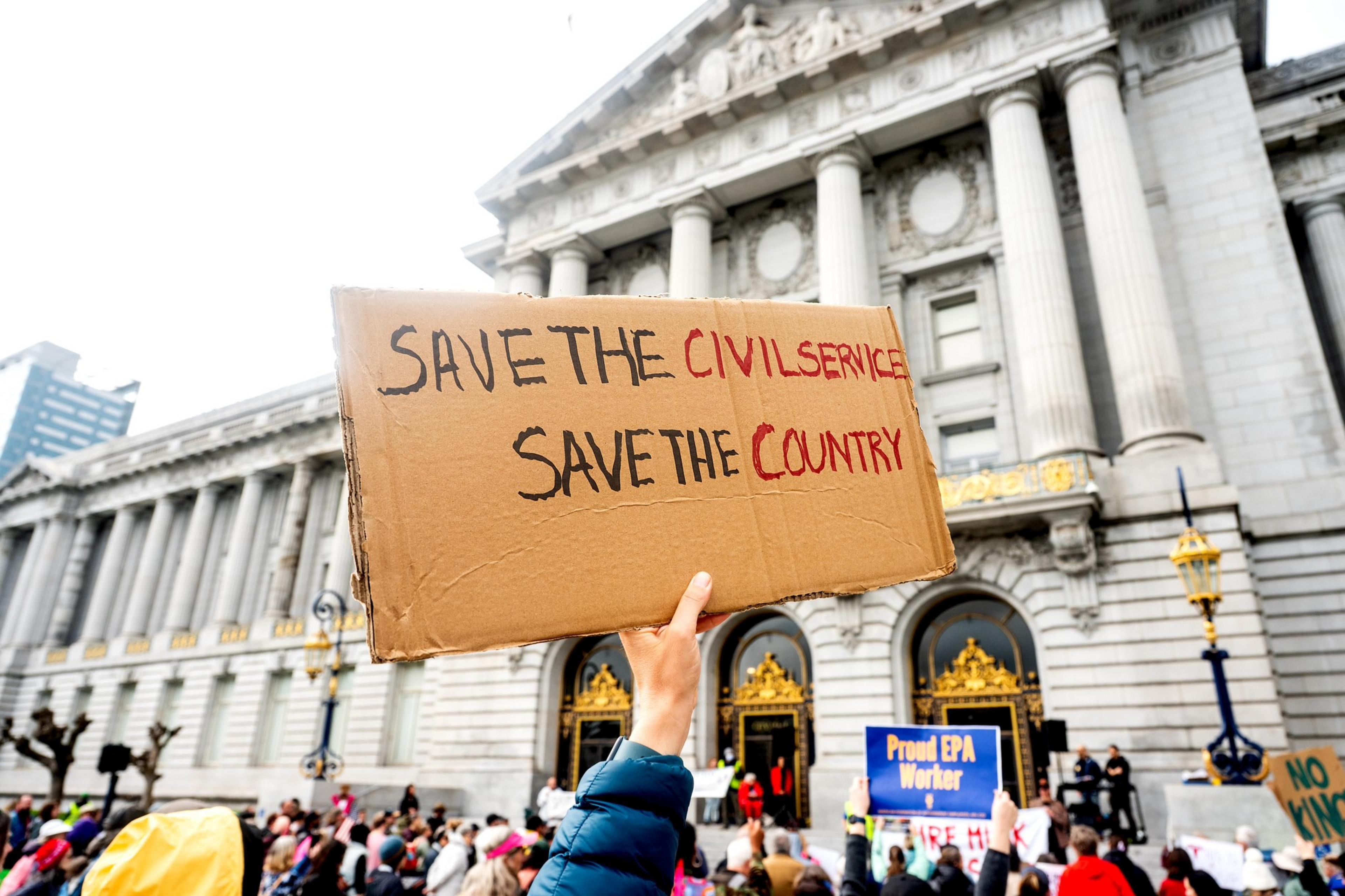 A hand holds a sign reading &quot;Save the Civil Service, Save the Country&quot; in front of a grand building with columns, while a crowd gathers below.
