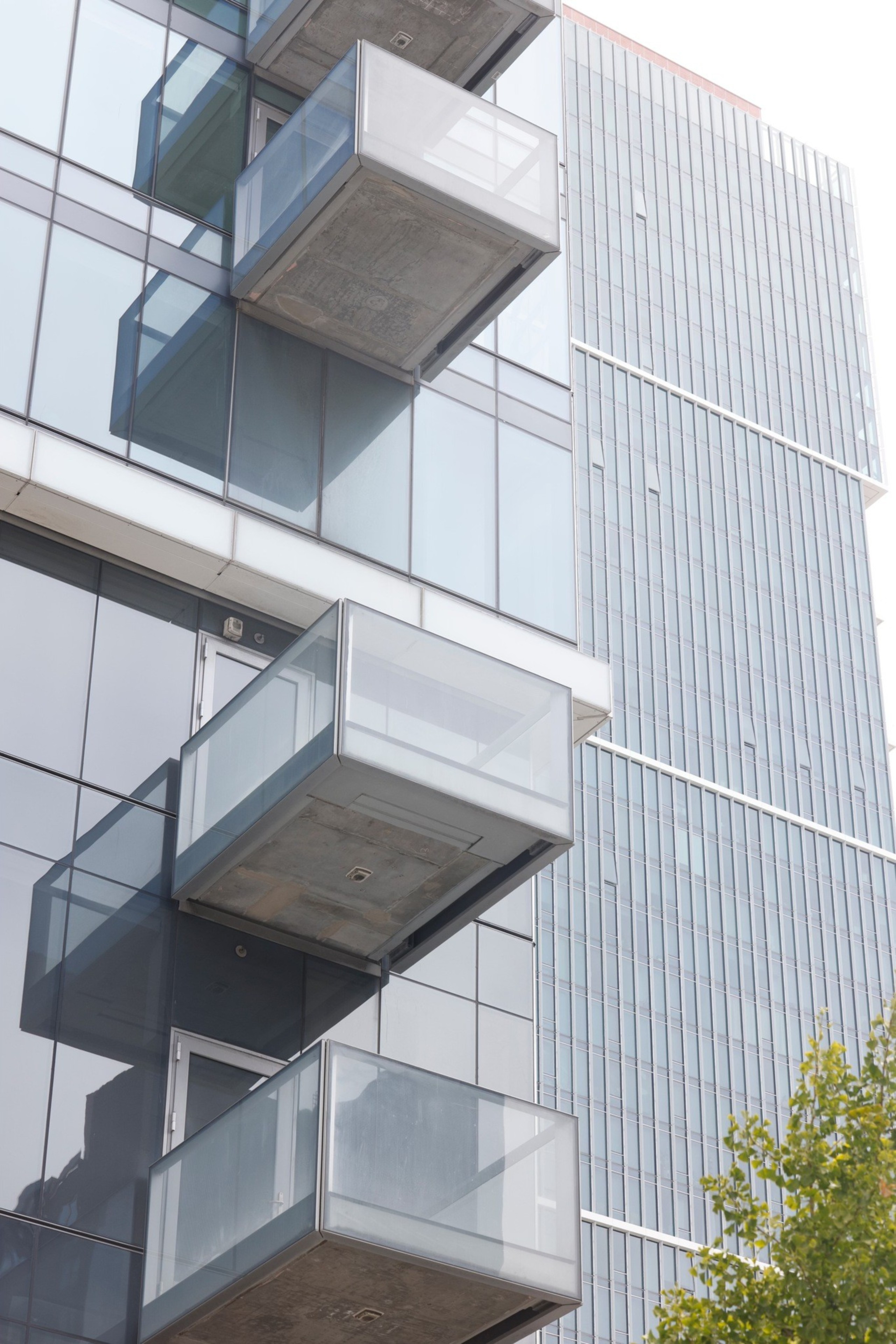 The image shows the side of a modern glass building with several jutting rectangular balconies. Another tall building with vertical lines is seen in the background.