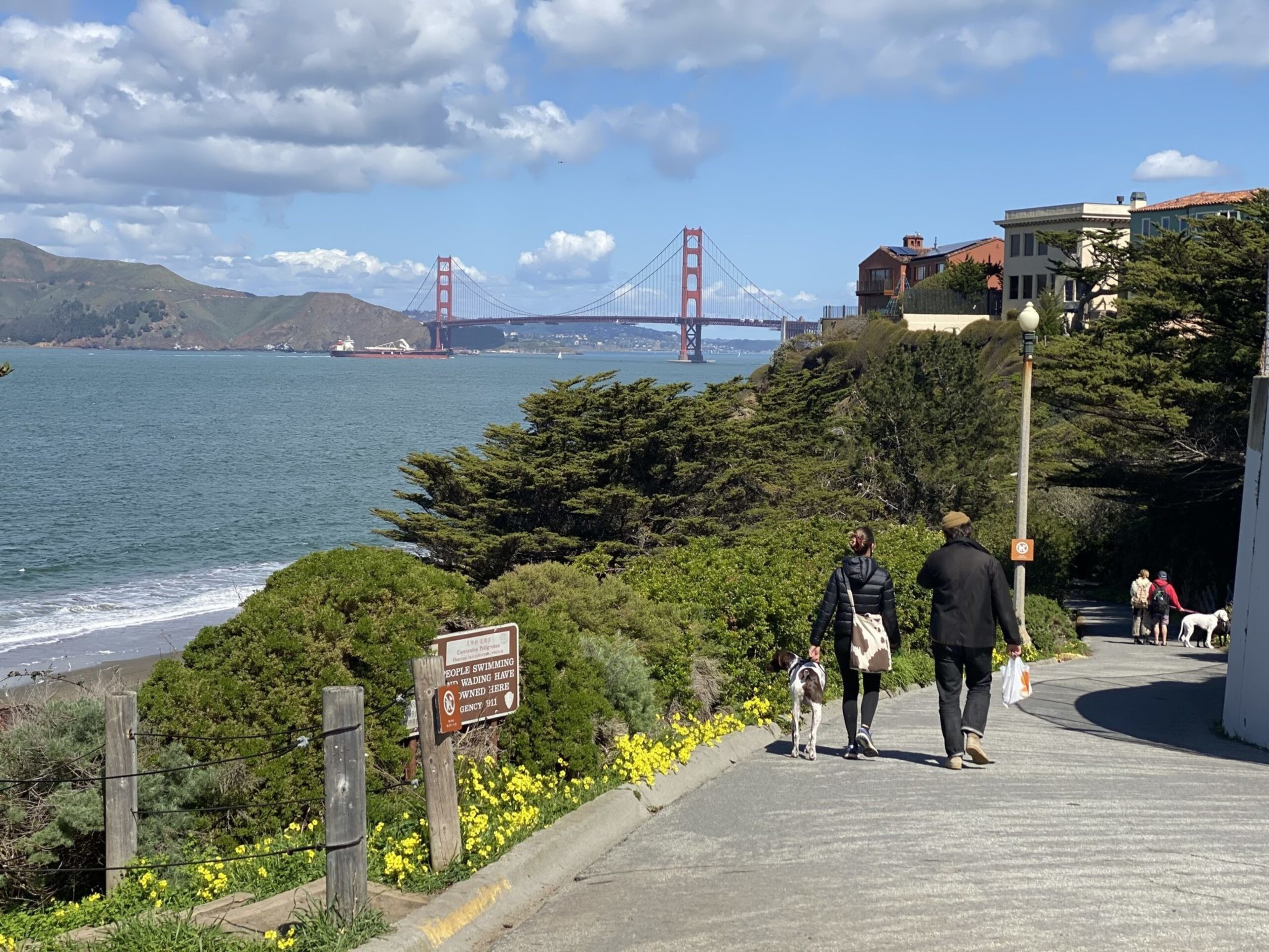 A coastal path with people walking dogs and the Golden Gate Bridge spanning blue water under a partly cloudy sky in the background.