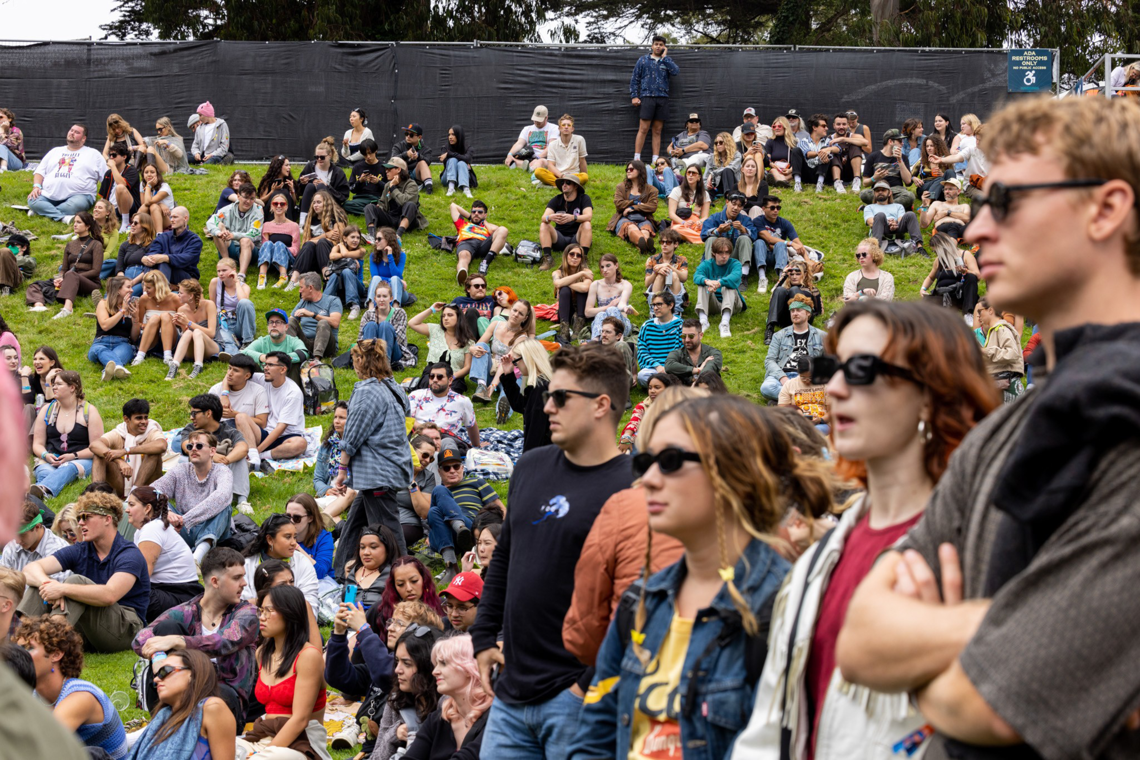 A large, diverse crowd of mostly young adults sits and stands on a grassy hill, many wearing sunglasses and casual clothing on a bright day.