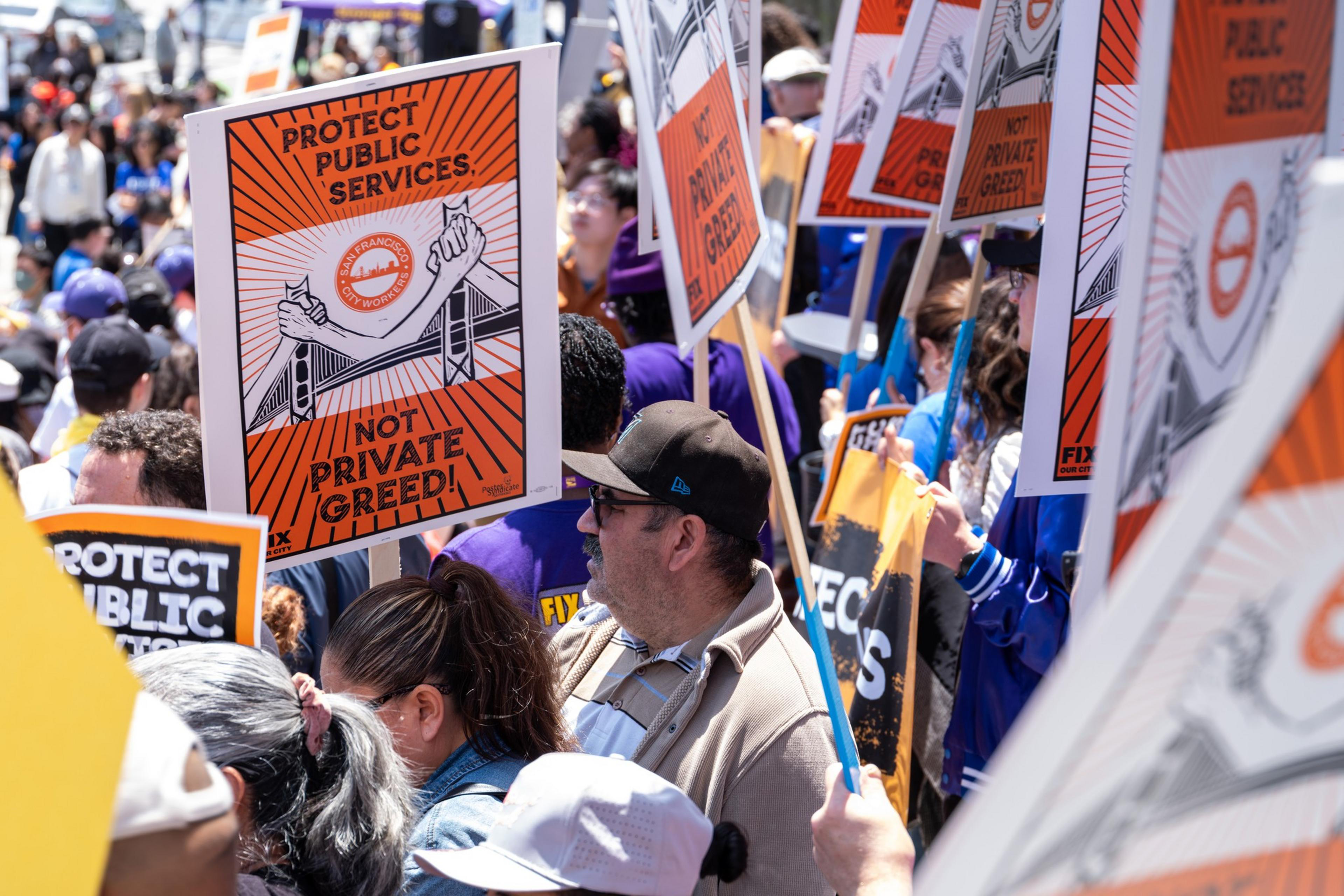The image shows a crowd of people holding signs during a public demonstration. The signs advocate for protecting public services and oppose private greed.